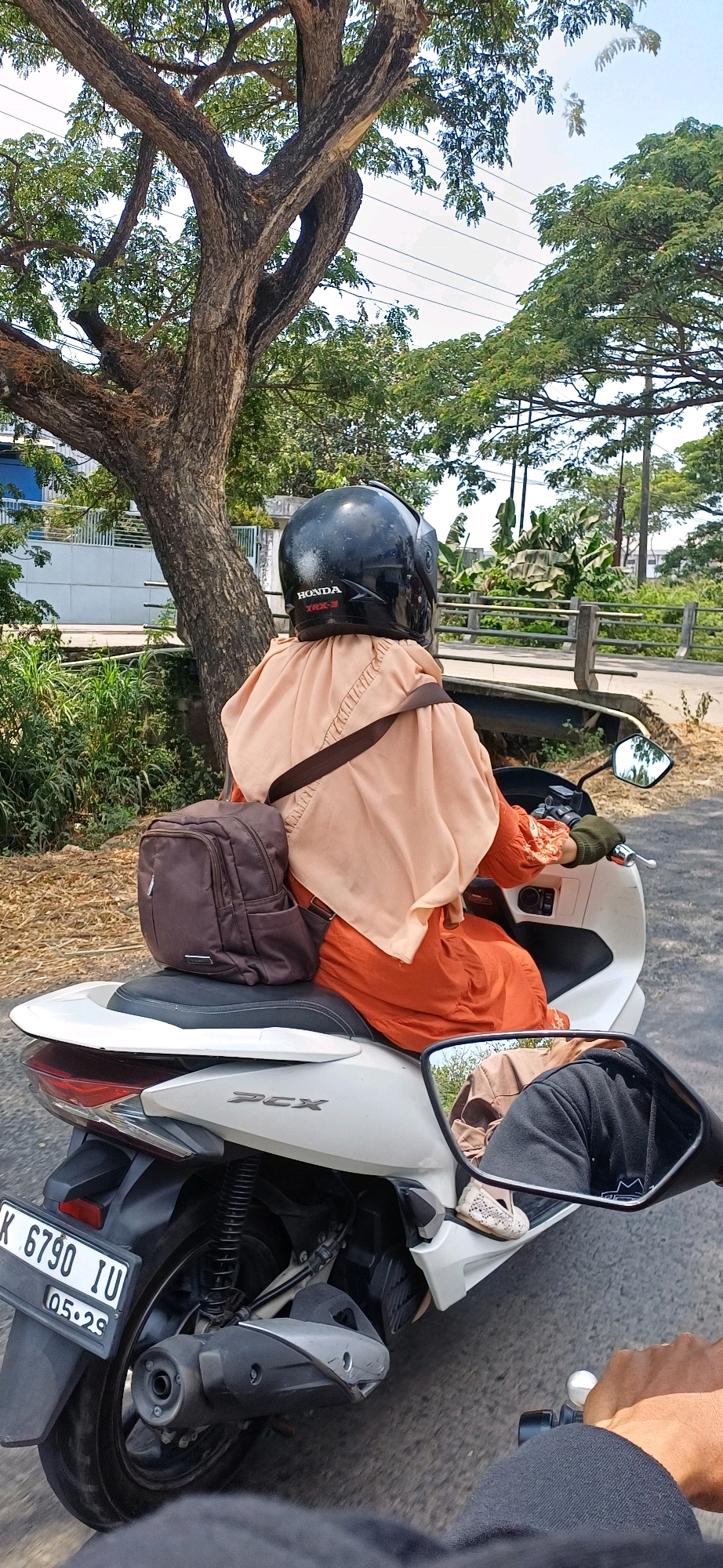 A person wearing a black helmet and an orange dress is riding a white motorcycle. The person has a brown backpack on their shoulder. The motorcycle is on a road with trees and vegetation on either side. The license plate on the motorcycle reads "K 6790 IU 05.29". In the reflection of the side mirror, another person wearing a black hoodie is visible. This scene appears to be in Kecamatan Pati, Indonesia, as indicated by the license plate.