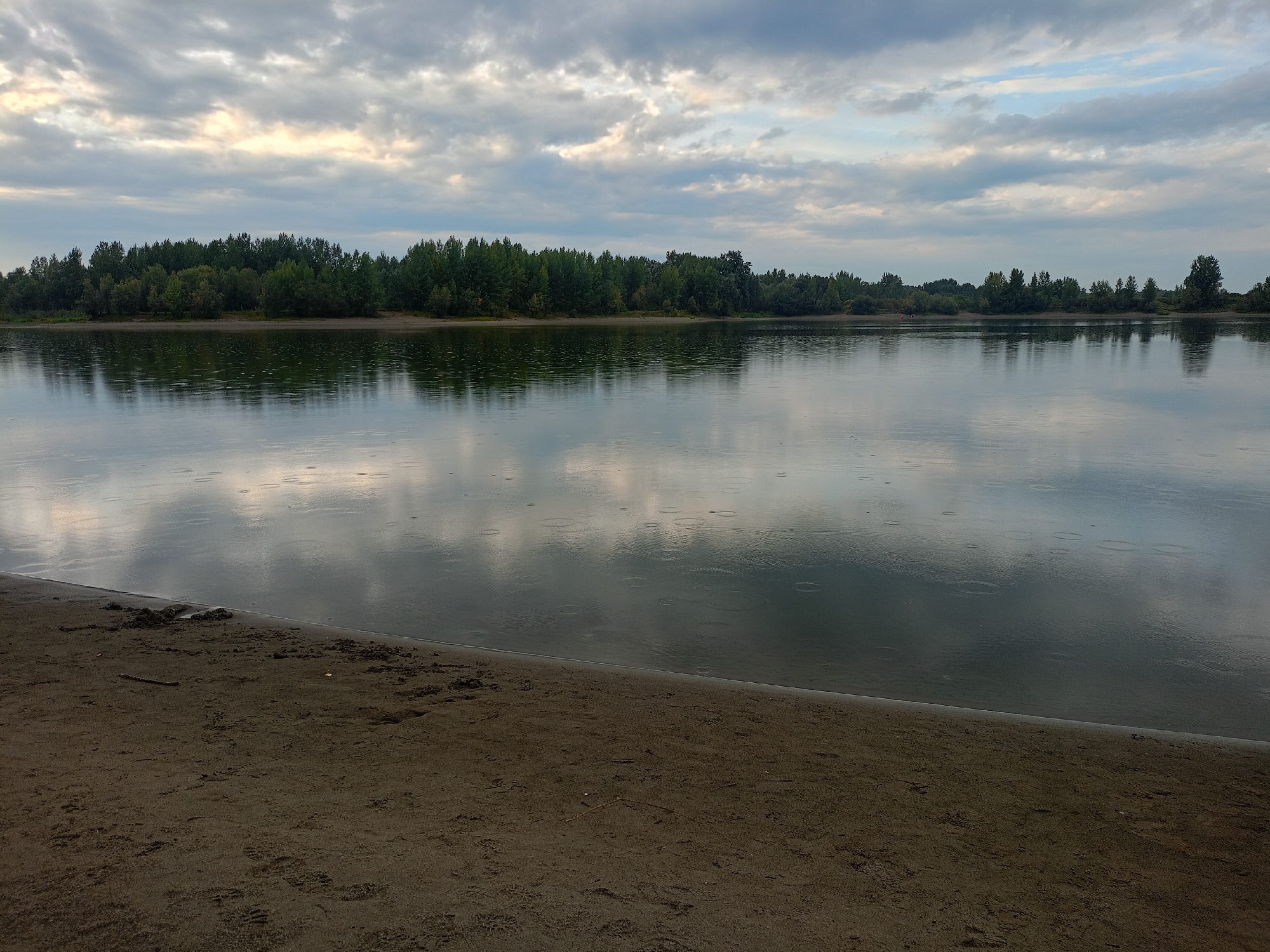 The image depicts a wide, calm river with a sandy embankment in the foreground. The sky is overcast with scattered clouds, allowing some sunlight to break through. The river reflects the trees on the opposite bank and the sky above. There are visible ripples on the water's surface, suggesting light rainfall. The vegetation on the far bank is dense and appears to be a mix of deciduous and coniferous trees. The sandy area in the foreground shows some texture and a few scattered pieces of debris, including a small stick. The overall scene is tranquil and natural, possibly a park or a natural reserve within or on the outskirts of Barnaul, Russia, given the presence of a large river.
