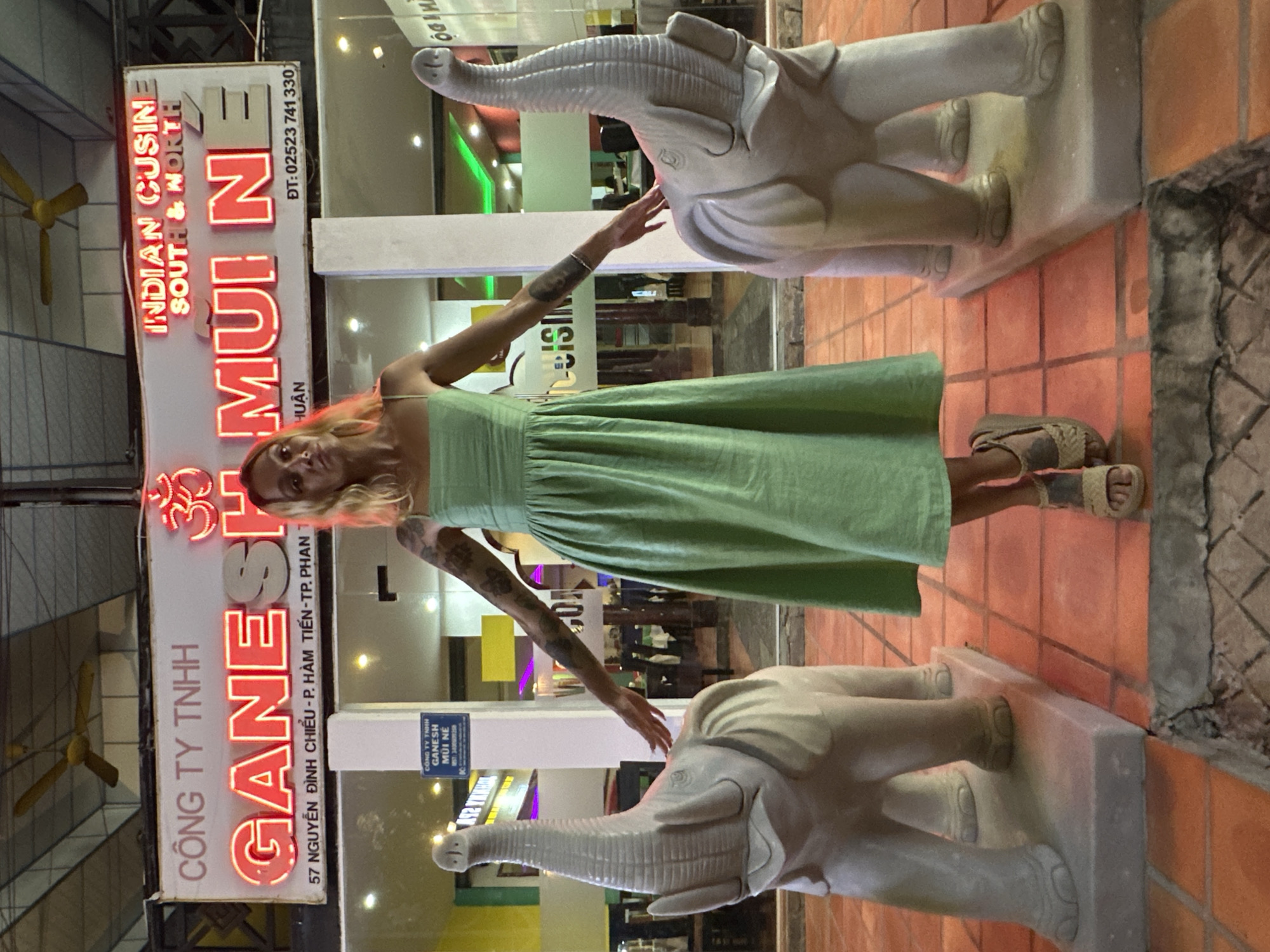 A woman with blonde hair and tattoos stands between two large elephant statues in front of a restaurant. The restaurant's sign reads "GANESH MUI NE" in neon letters, with "INDIAN CUISINE SOUTH & NORTH" above it. The address "57 NGUYEN DINH CHIEU - P. HAM TIEN - TP. PHAN THIET" is also visible, indicating the location is in Phu Thuy Ward, Vietnam. The woman is wearing a light green sleeveless dress and tan sandals, with her arms outstretched. The elephant statues are made of stone or concrete and are positioned on either side of her. The ground is tiled with orange tiles. The lighting suggests it is evening or nighttime.