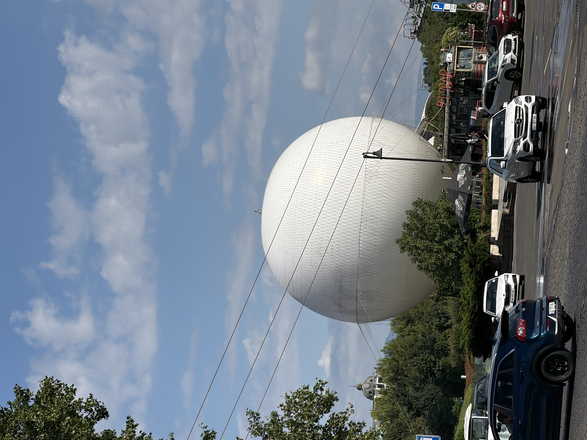 In Tbilisi, Georgia, this photo captures a sunlit day with a large, white, spherical structure dominating the scene. The structure is a modern concert hall, known as the Tbilisi Concert Hall or The Orb. It's surrounded by lush green trees and foliage. In the foreground, a traffic intersection is visible with several cars driving or parked on a cobblestone road. Among the vehicles is a dark blue Kia SUV, a white Mercedes-Benz SUV, and a white Skoda sedan. A red SUV is parked on the right side of the frame. Behind the spherical structure, a vintage-style food truck with a neon sign that reads "24 HR BAR" is parked, along with outdoor seating areas and umbrellas. The sky is a vibrant blue with scattered white clouds, and overhead power lines crisscross the frame. In the distance, a domed building and some residential structures can be seen amongst the trees, hinting at the urban landscape of Tbilisi.