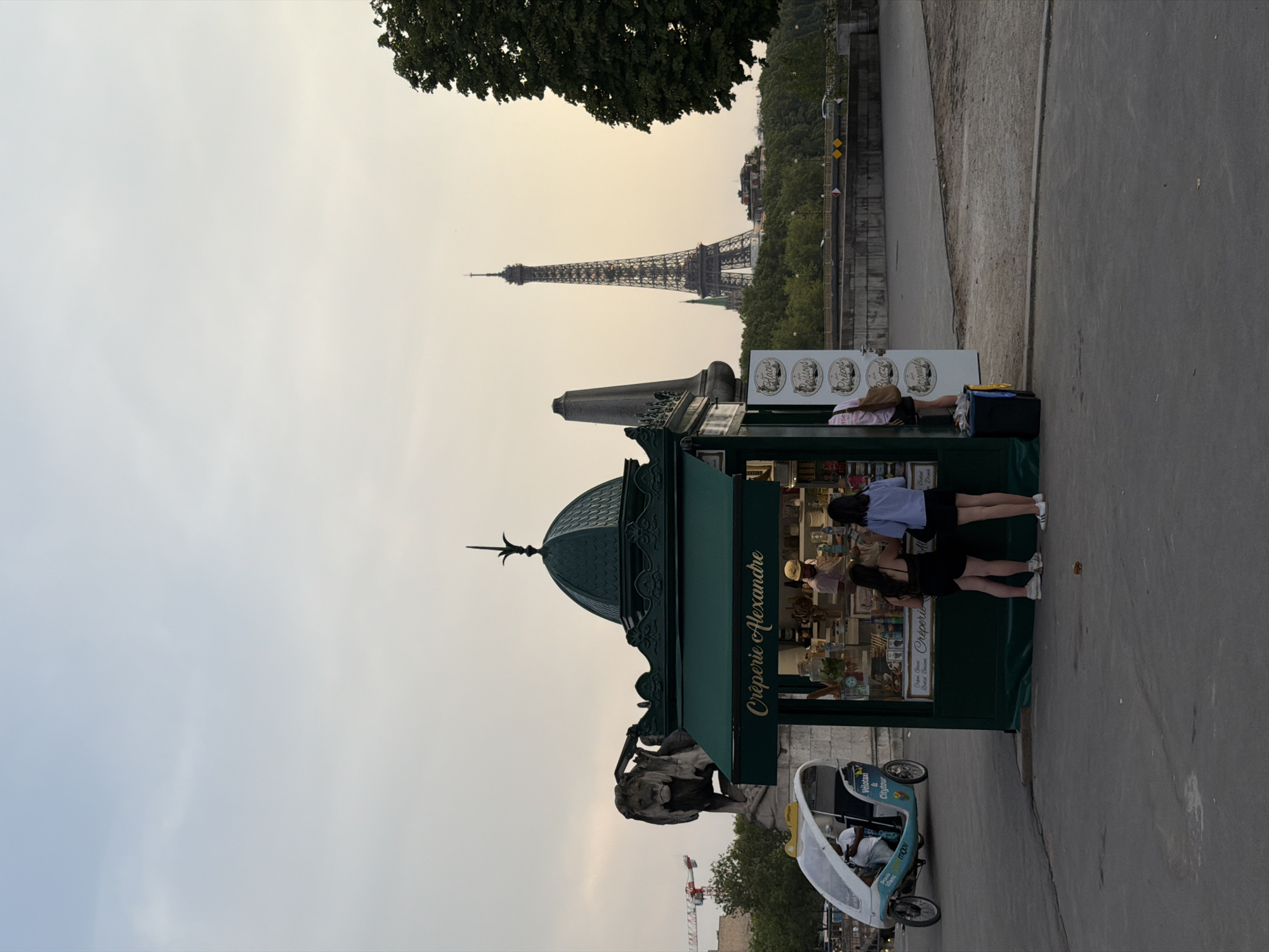 The image shows a creperie stand on a street in Paris, France, with the Eiffel Tower visible in the background. The creperie stand is green and has a sign that reads "Crêperie Alexandre". There are two women standing in front of the stand, looking at the menu. A man wearing a yellow hat is working behind the counter. To the left of the creperie stand, there is a three-wheeled bicycle taxi. In the background, a large stone lion statue is visible, as well as a construction crane. The sky is overcast, and the lighting suggests it is either early morning or late afternoon.