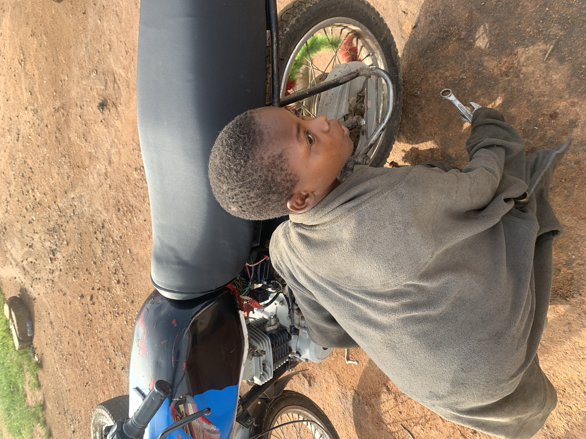 An overhead shot captures a young child intently working on a motorcycle outdoors on a dry, reddish-brown dirt ground. The child, appearing to be a boy of African descent with short, dark hair, is dressed in a long-sleeved, grey fleece-like top. He is crouched beside the front portion of a black and blue motorcycle, focusing on the area near its front wheel and engine.
In his right hand, the child holds a silver-colored wrench, indicating an activity of repair or maintenance. His profile shows a focused or curious expression as he looks at the motorcycle's components. The motorcycle itself appears well-used, with scratches visible on its bodywork and exposed engine parts. It features a black seat and spoked wheels.
The scene is brightly lit by natural sunlight, suggesting a clear, dry day, likely midday or early afternoon, with distinct shadows visible on the ground. The unpaved, dusty earth points to a rural or semi-urban setting, consistent with the Barakin Lerawi, Nigeria context. There is no visible text within the image.