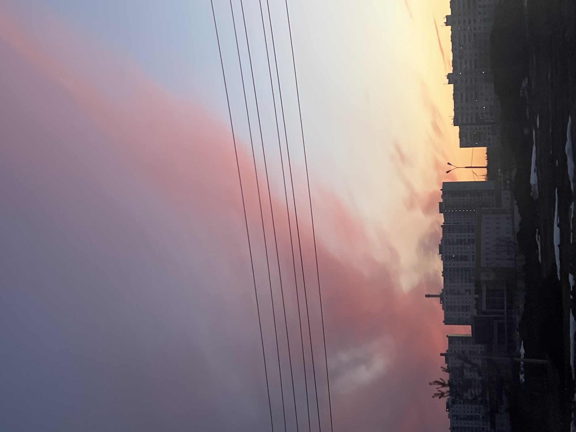 The image shows a cityscape at dusk or dawn, possibly in Usolye, Russia, based on the urban environment. The sky transitions from a pale blue to hues of orange and pink, with streaky clouds reflecting the light. Overhead electrical wires run across the sky, dividing the open space from the built environment. The buildings are mid-rise to high-rise residential structures. The foreground is dark, suggesting land with patches of what appears to be snow. There are no people visible in the image.