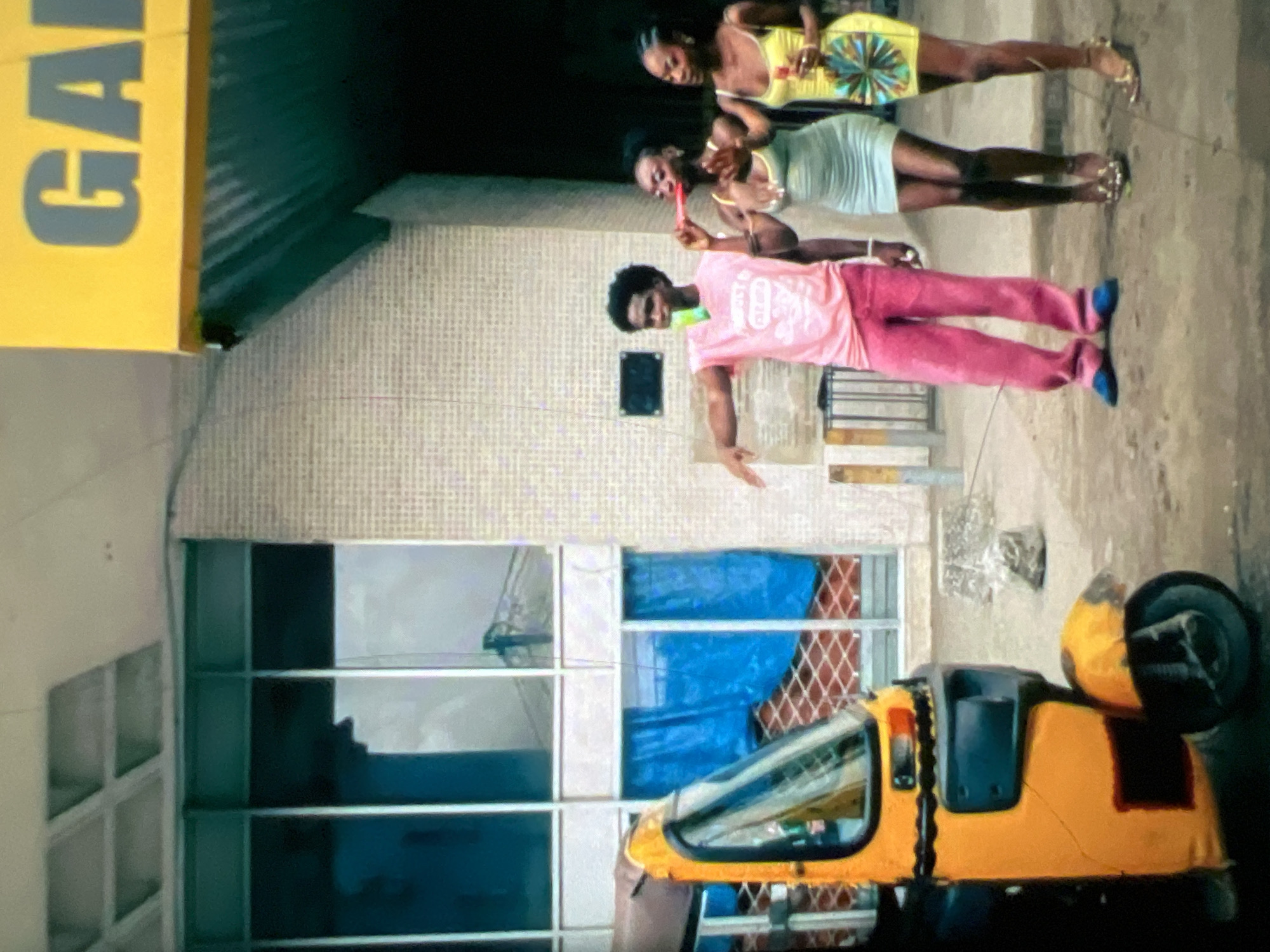 In Lagos, Nigeria, a man wearing a pink sleeveless shirt and pink pants stands with a green popsicle in his hand, facing three women. The woman to his left wears a light blue dress and holds a red popsicle. The woman to her left wears a yellow dress and holds a yellow popsicle and a colorful purse. They are standing on a street in front of a building with a yellow sign that reads "GA". A yellow auto-rickshaw is parked to the left of the frame. The ground is made of dirt and debris.