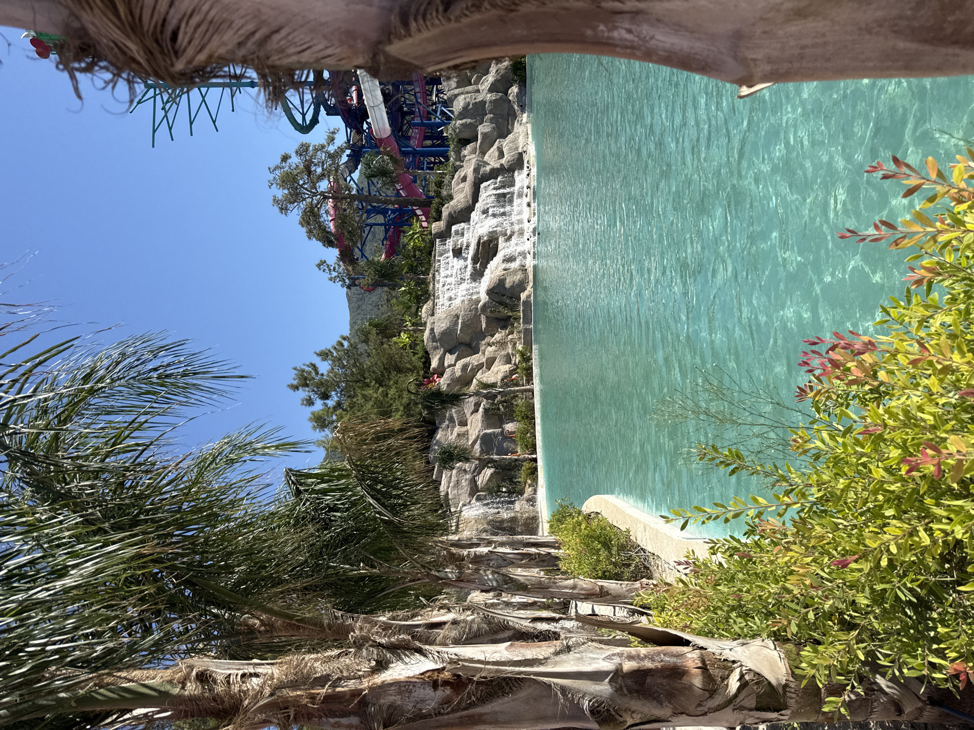 The image depicts a serene view of a resort pool area with a waterfall cascading down a rocky embankment. Palm trees frame the scene, with lush green foliage and vibrant red-tipped leaves in the foreground. The turquoise water of the pool is clear and inviting, reflecting the bright blue sky. In the background, a water park with colorful slides and structures is visible, hinting at the leisure and entertainment offerings of the location. The overall atmosphere is one of tropical relaxation and fun. This scene is likely from a resort or water park in Bodrum, Turkey, a popular tourist destination known for its beautiful beaches and vibrant atmosphere.