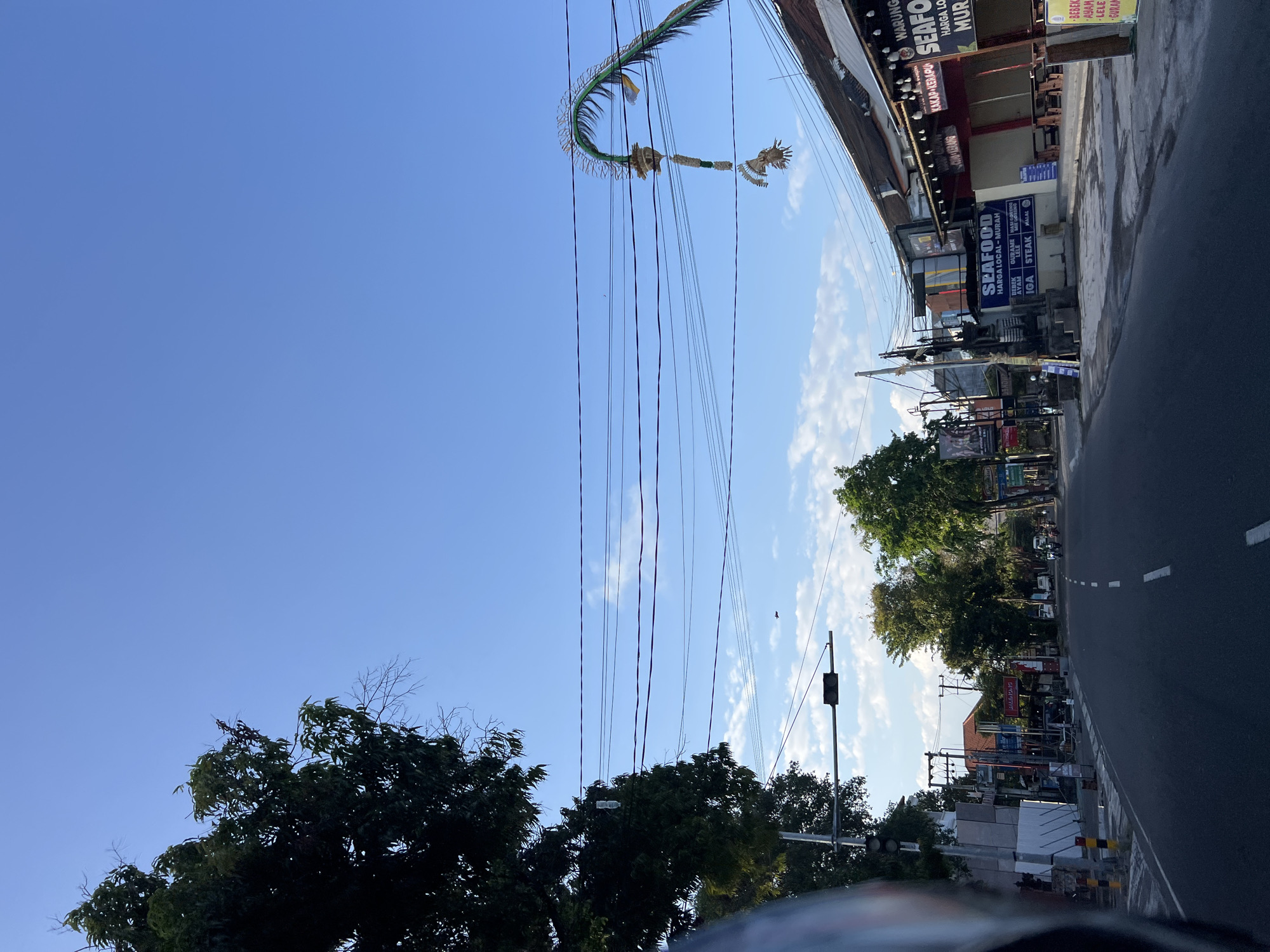 The image shows a street scene in Kuta, Indonesia. The road is paved and has white dashed lines marking the lanes. On the left side of the road, there are trees, a traffic light, and buildings. On the right side of the road, there are more trees and buildings, including a seafood restaurant with outdoor seating. There are also power lines crisscrossing the sky. The sky is blue with a few scattered clouds. A decorative green archway with hanging ornaments is visible in the upper right portion of the image. A few motorbikes are visible on the road. The overall impression is of a typical street in a tourist area.