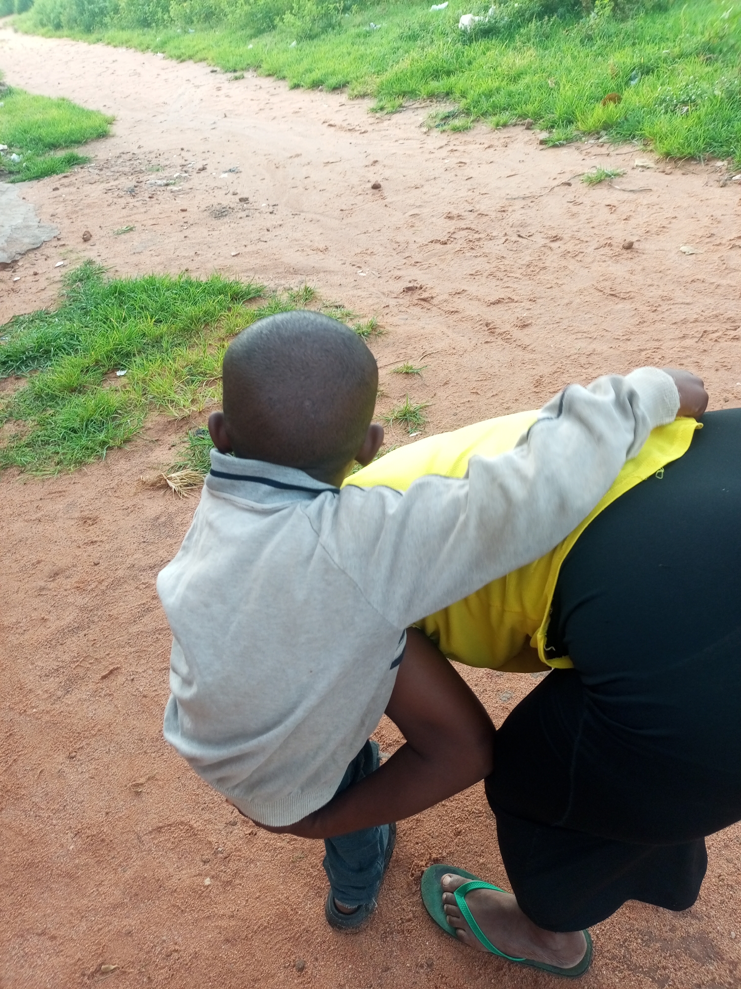 A young person in a grey sweater and blue jeans is bending over and hugging a person wearing a yellow top and black pants. The younger person's head is visible from behind. They are on a dirt path lined with green grass.