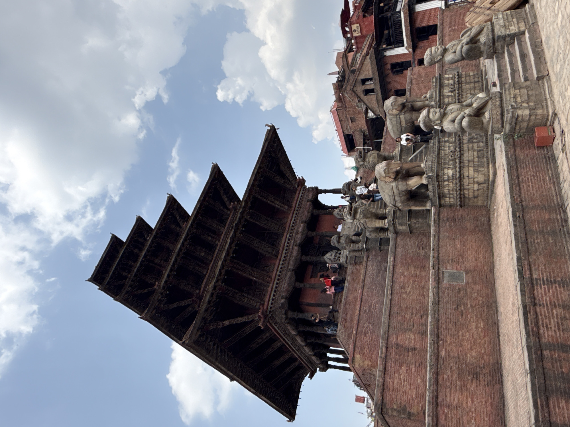 The image captures the five-story Nyatapola Temple in Bhaktapur, Nepal, under a bright sky with scattered clouds. The temple's tiered roofs rise sharply against the sky, with dark wooden beams supporting each level. The temple stands on a brick base, and stone sculptures of figures are positioned at various levels of the staircase leading up to the temple. People are visible on and around the stairs of the temple. Other brick buildings are visible around the Nyatapola Temple.