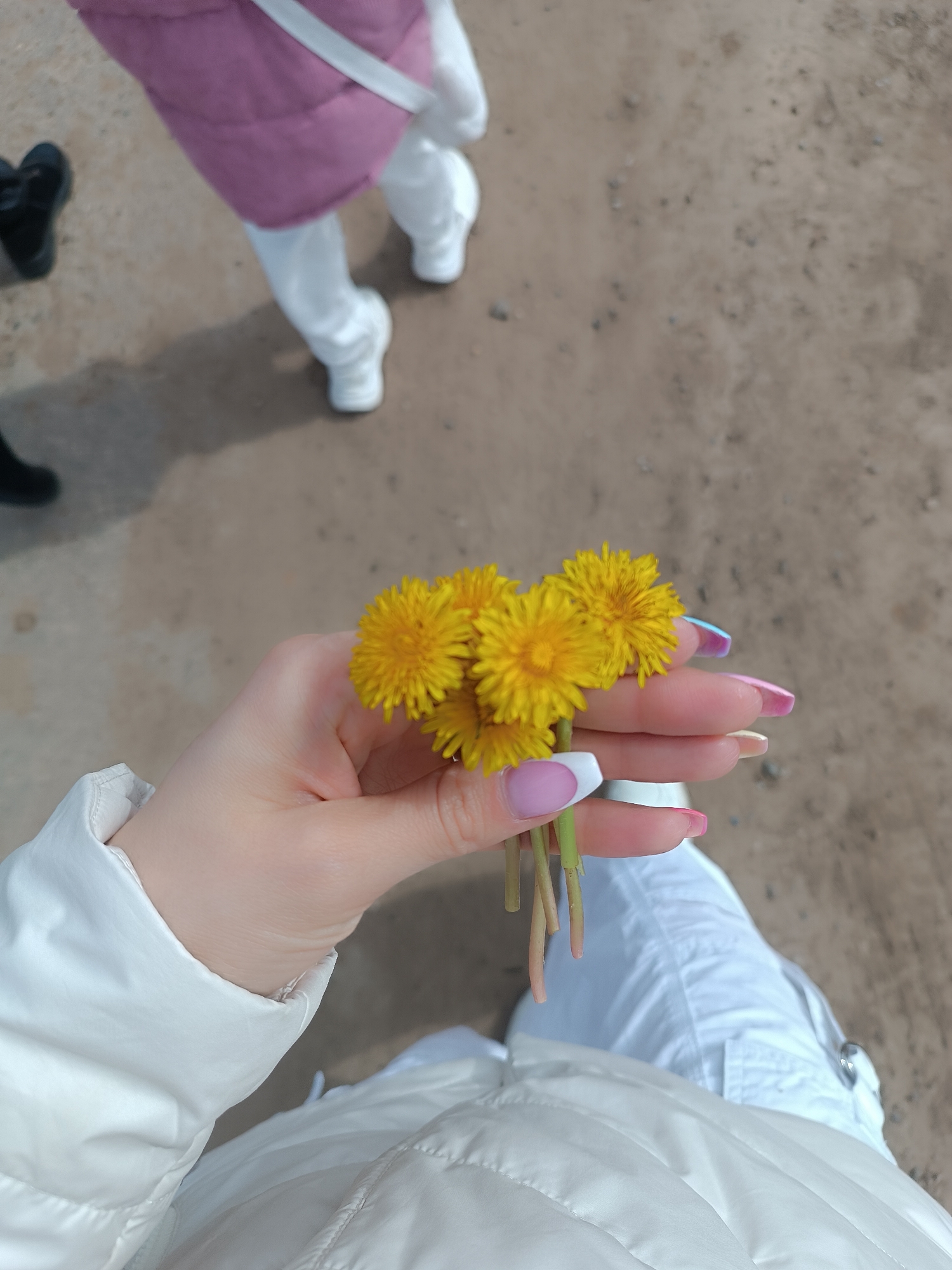 A first-person, low-angle shot reveals a hand holding a small bouquet of yellow dandelions. The person's nails are manicured with a mix of French tips, pink, and iridescent designs. The person is wearing a white quilted jacket and white pants. In the background, other people are walking on a sandy or gravelly surface. One person is wearing a pink quilted jacket and white pants. Another person's black shoes are visible. The photograph appears to be taken outdoors, possibly in a park or recreation area. The location is Chaikovsky, Russia.