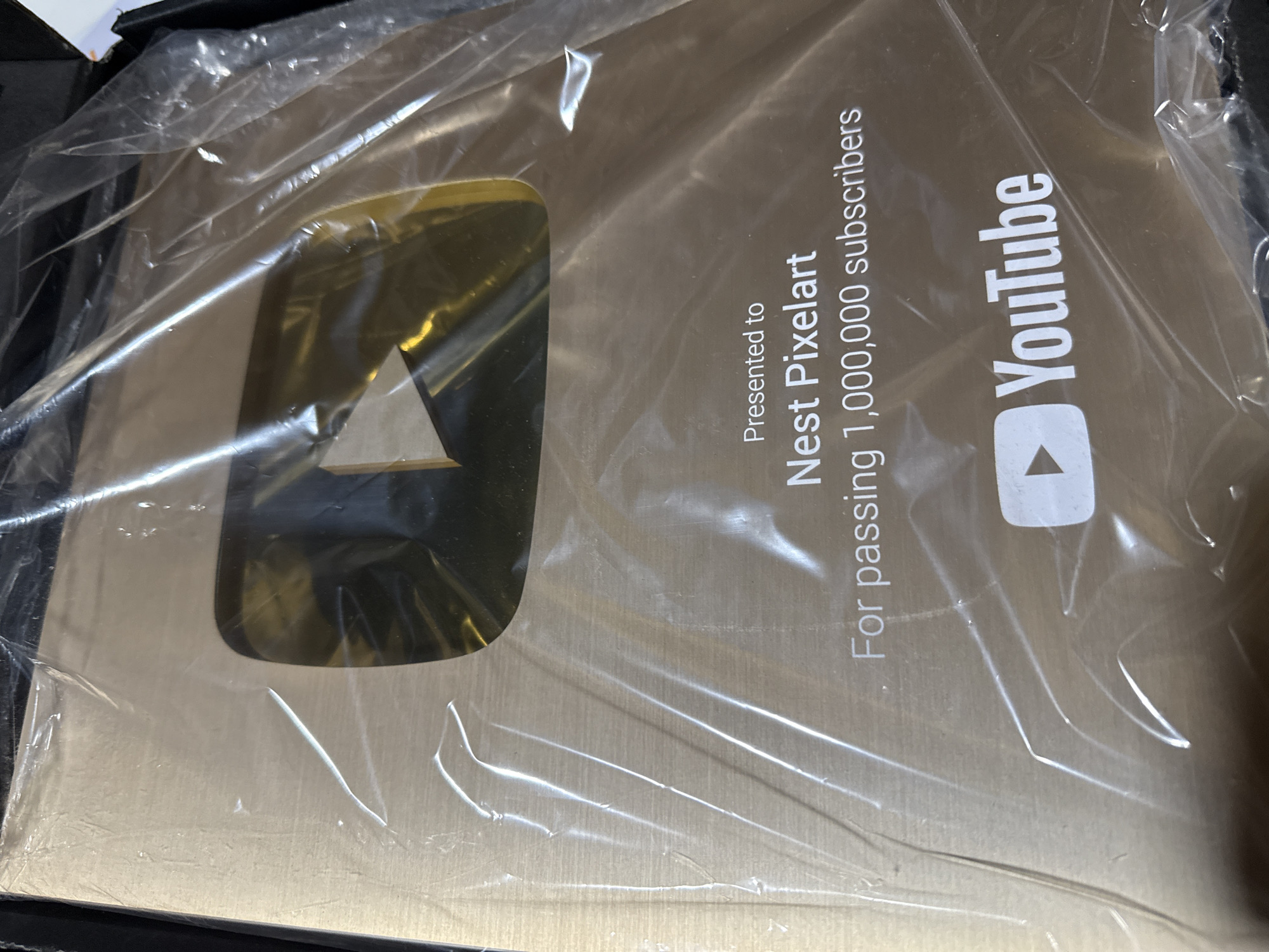 A YouTube Gold Play Button award, still inside its plastic packaging, is the central focus of the image. The award is presented to "Nest Pixelart" for surpassing 1,000,000 subscribers. The YouTube logo is prominently displayed. The location, Puebla, Mexico, is not evident from the image. The surrounding environment is obscured by the packaging and the dark edges of what appears to be a box.