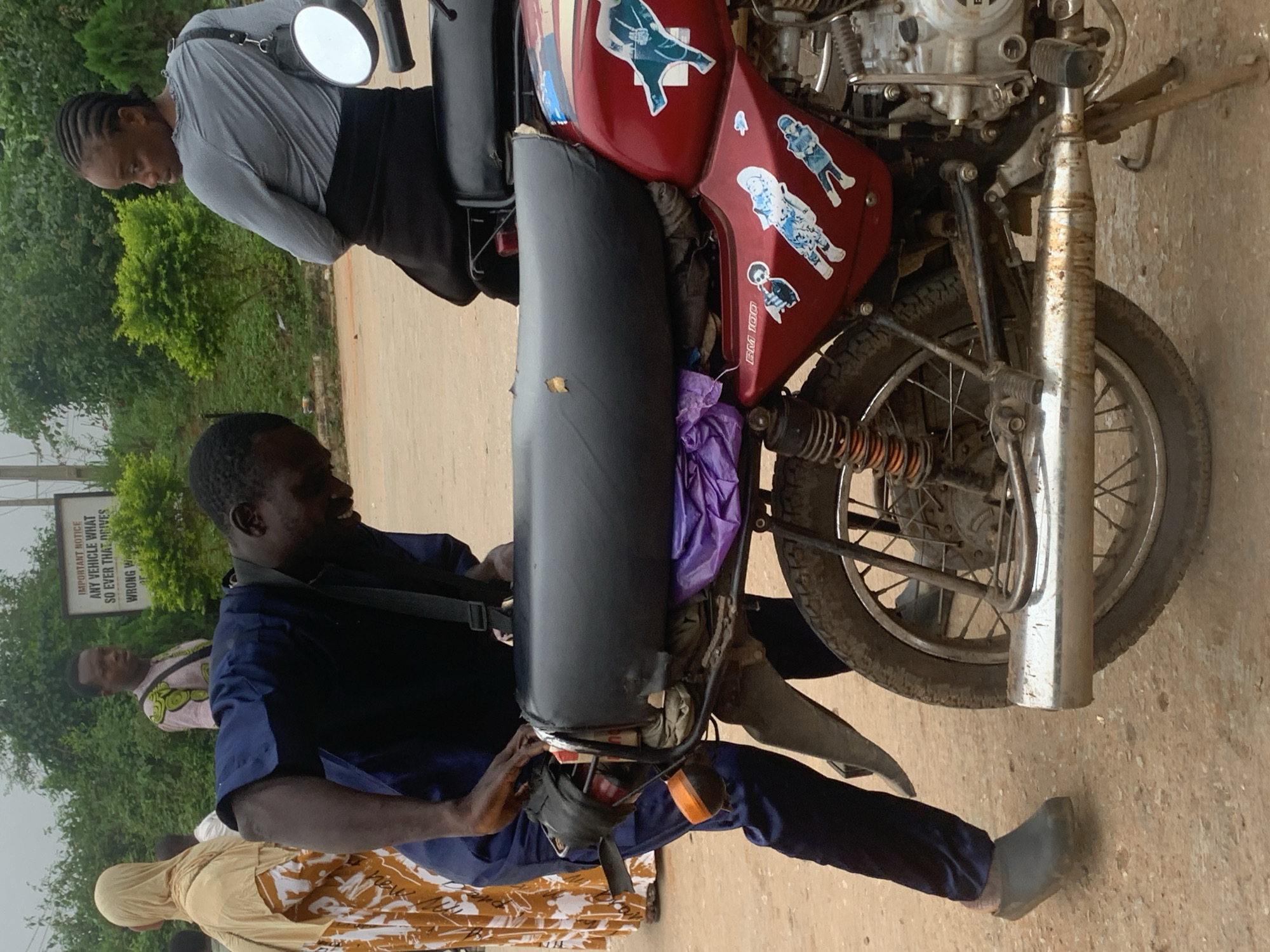 In Shagamu, Nigeria, a man in a navy blue uniform with a black strap across his chest is adjusting the cargo on the back of a red motorcycle. The motorcycle has a black seat with a small tear, and is adorned with several blue and white stickers. A purple plastic bag is tied to the motorcycle rack. To the left of the man, a woman wearing a light brown hijab and an orange and white patterned dress stands with her back to the camera. Further in the background, a man in a pink and green patterned shirt is visible, along with a sign that reads "IMPORTANT NOTICE ANY VEHICLE THAT SO EVER THAT DRIVES WRONG W". A woman with braided hair wearing a grey long-sleeved shirt and a black skirt is seated on the motorcycle, looking away from the man. The ground is dusty and unpaved, with some green foliage in the background.