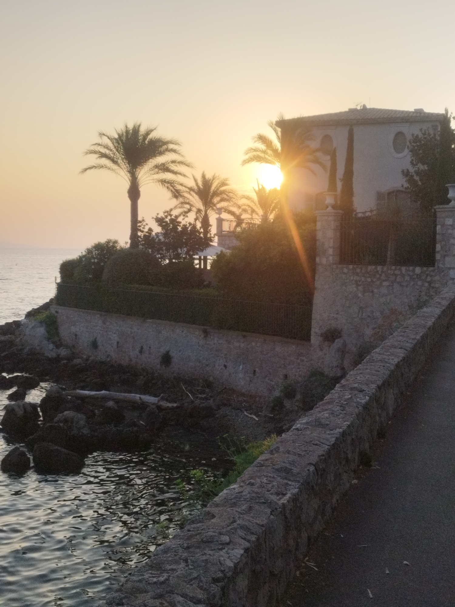 The image depicts a coastal scene in Antibes, France, during a sunset. In the foreground, a paved pathway runs alongside a rustic stone wall that borders a rocky shoreline. The water of the Mediterranean Sea is visible on the left, with gentle waves lapping against the dark rocks.
In the midground, the stone wall continues, topped with a dense hedge. Behind the hedge, a white villa with a tiled roof is visible, partially obscured by palm trees and other vegetation. The sun is setting behind the villa, casting a warm glow and creating sun flares that extend across the scene. Two tall, slender cypress trees stand near the villa.
The sky is a soft, hazy orange and yellow, characteristic of a sunset. The overall atmosphere is serene and picturesque. There are no people or animals visible in the image. The scene is a typical coastal landscape found in the region.