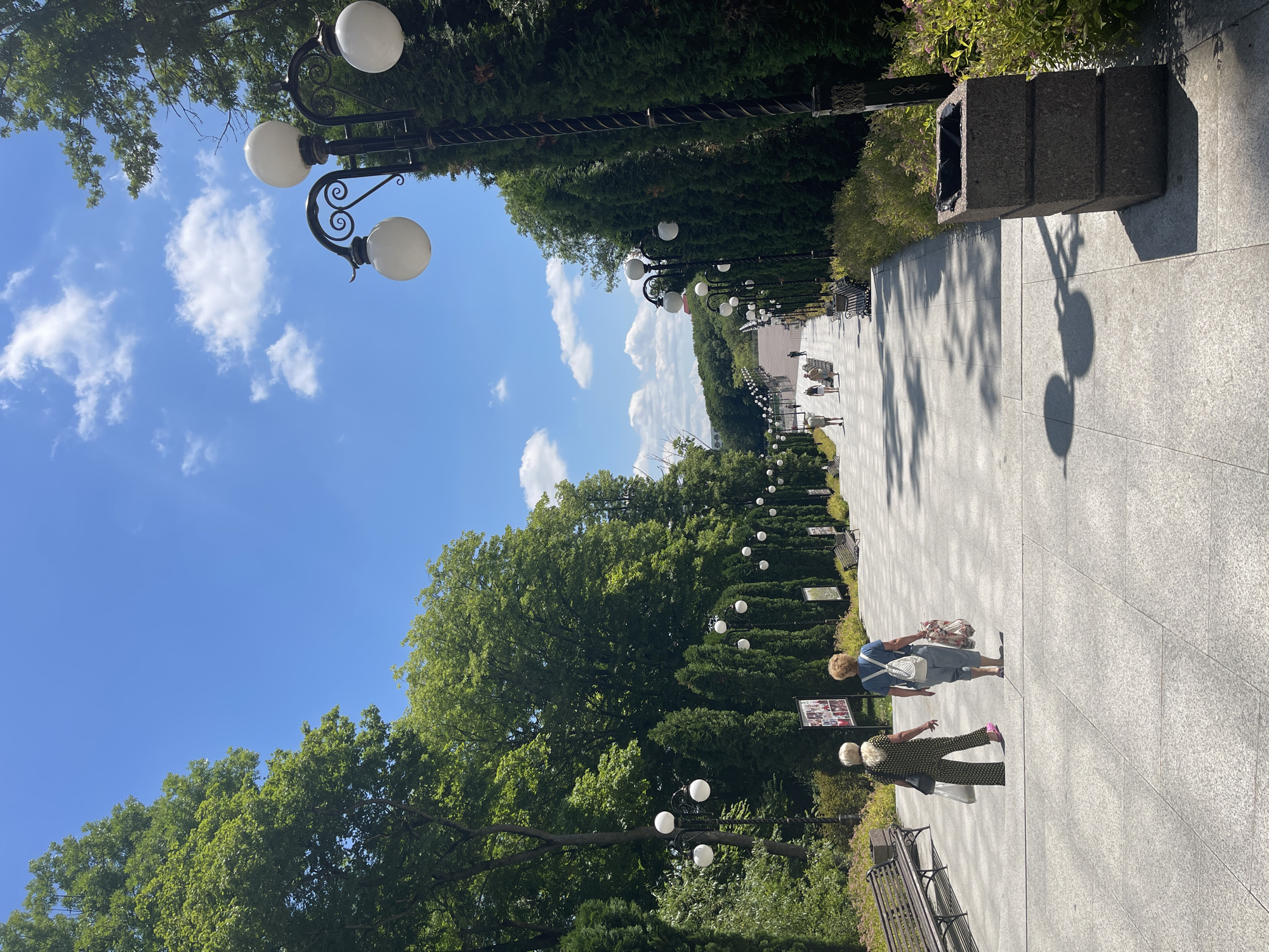 This image shows a paved walkway lined with trees and lampposts in Zheleznovodsk, Russia. The walkway is wide and made of light-colored stone tiles. On the left side of the walkway, there are tall, mature trees and manicured evergreen bushes. Decorative lampposts with multiple white globe lights are spaced evenly along the path. On the right side of the walkway, there are more evergreen bushes, benches, and a large granite trash receptacle.
Two women are walking on the walkway. One woman in a blue shirt and light-colored pants is walking away from the camera, carrying a white backpack and a patterned bag. Another woman in a patterned outfit is walking towards the camera.
The scene appears to be set during the day, indicated by the bright sunlight casting shadows on the walkway and the blue sky with scattered white clouds. The weather seems pleasant, suitable for outdoor activities. The overall atmosphere is peaceful and serene, suggesting a park or a public garden setting. No visible text is present in the image.