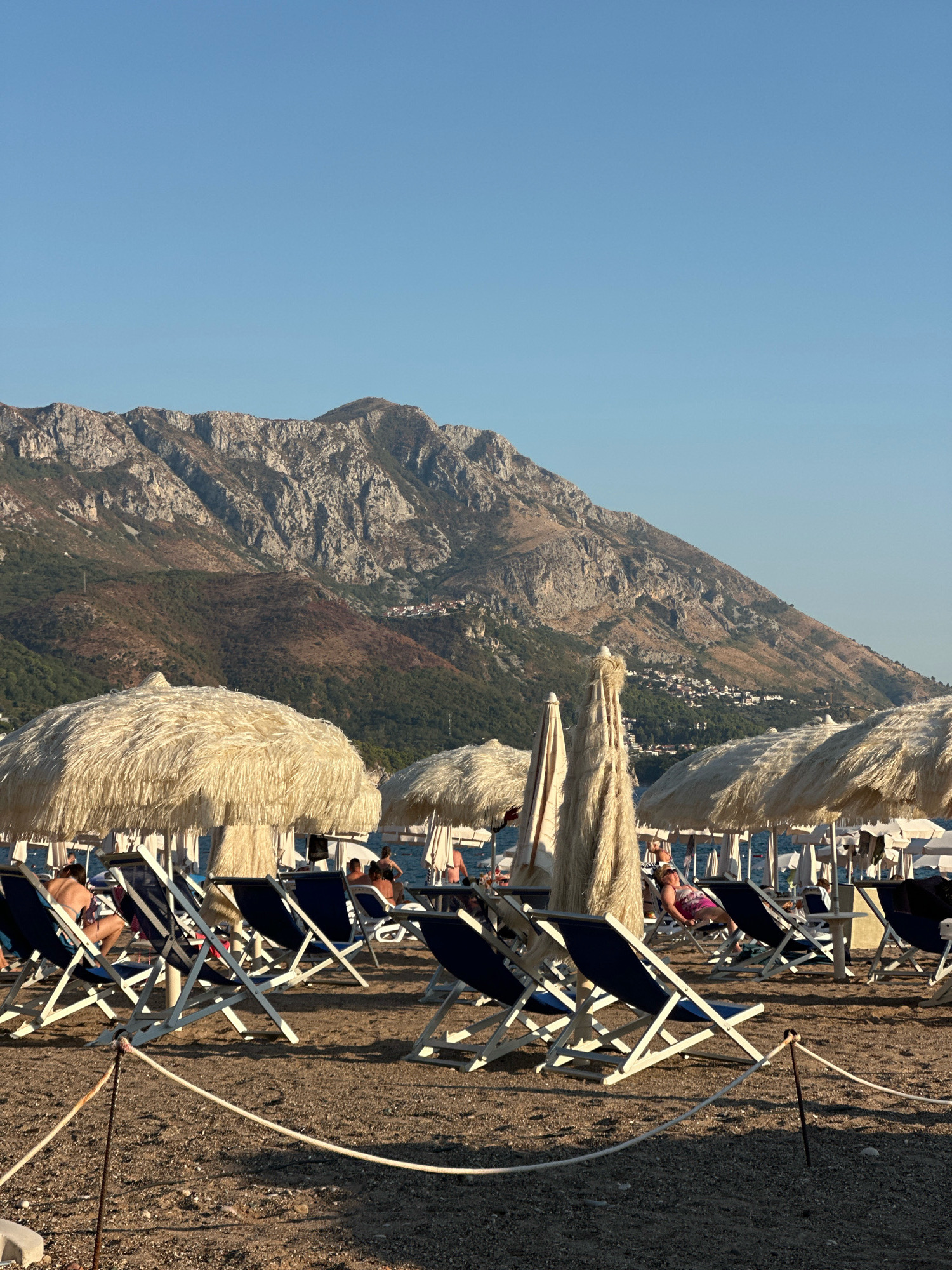 The image depicts a beach scene in Boreti, Montenegro. In the foreground, numerous beach chairs with blue canvas seating are arranged on a sandy and pebbly shore. Several straw-covered umbrellas are set up, providing shade over some of the chairs. A white rope barrier is visible in the lower portion of the frame, running horizontally across the beach. In the mid-ground, people are seated in the chairs, some appearing to be relaxed or sunbathing. In the background, a body of water, likely the Adriatic Sea, is visible, with a few boats or structures on it. Towering over the scene is a large, rugged mountain range, its slopes covered in a mix of vegetation and exposed rock. Small buildings are scattered on the lower parts of the mountain. The sky is clear and a bright blue. The lighting suggests it is late afternoon or early evening, with long shadows cast by the chairs and umbrellas.