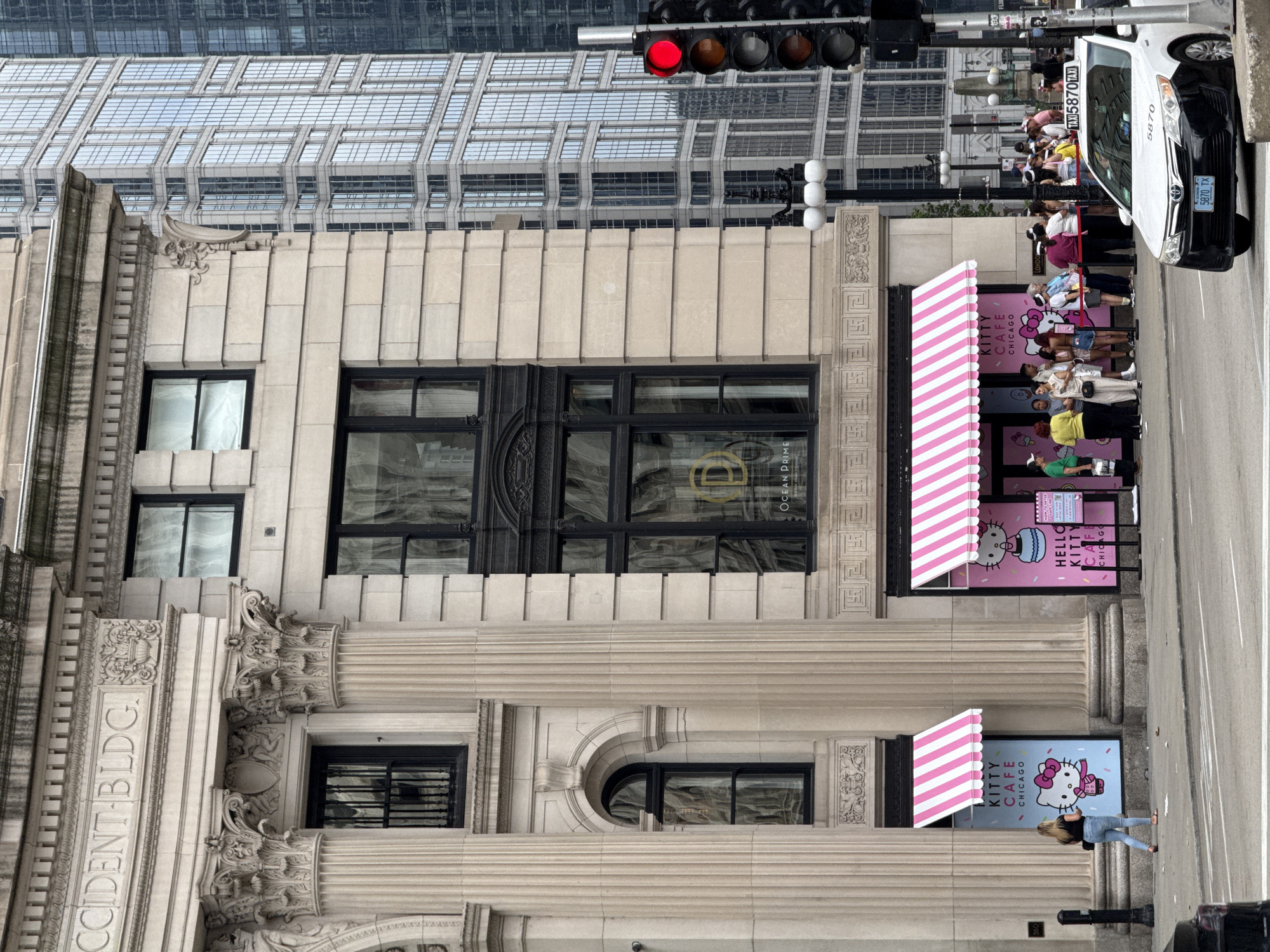 The image depicts the exterior of the Kitty Cafe in Chicago, USA. The cafe is located next to a grand stone building with classical architecture, featuring large columns and ornate carvings. The cafe itself is brightly decorated with pink and white striped awnings and large posters of Hello Kitty. A line of people is visible waiting to enter the cafe. In the foreground, a white taxi with the number 5870 is parked on the street. Traffic lights are visible to the right, showing a red light. The background shows modern glass skyscrapers, indicating an urban setting. The street is paved, and there is a sidewalk in front of the buildings.