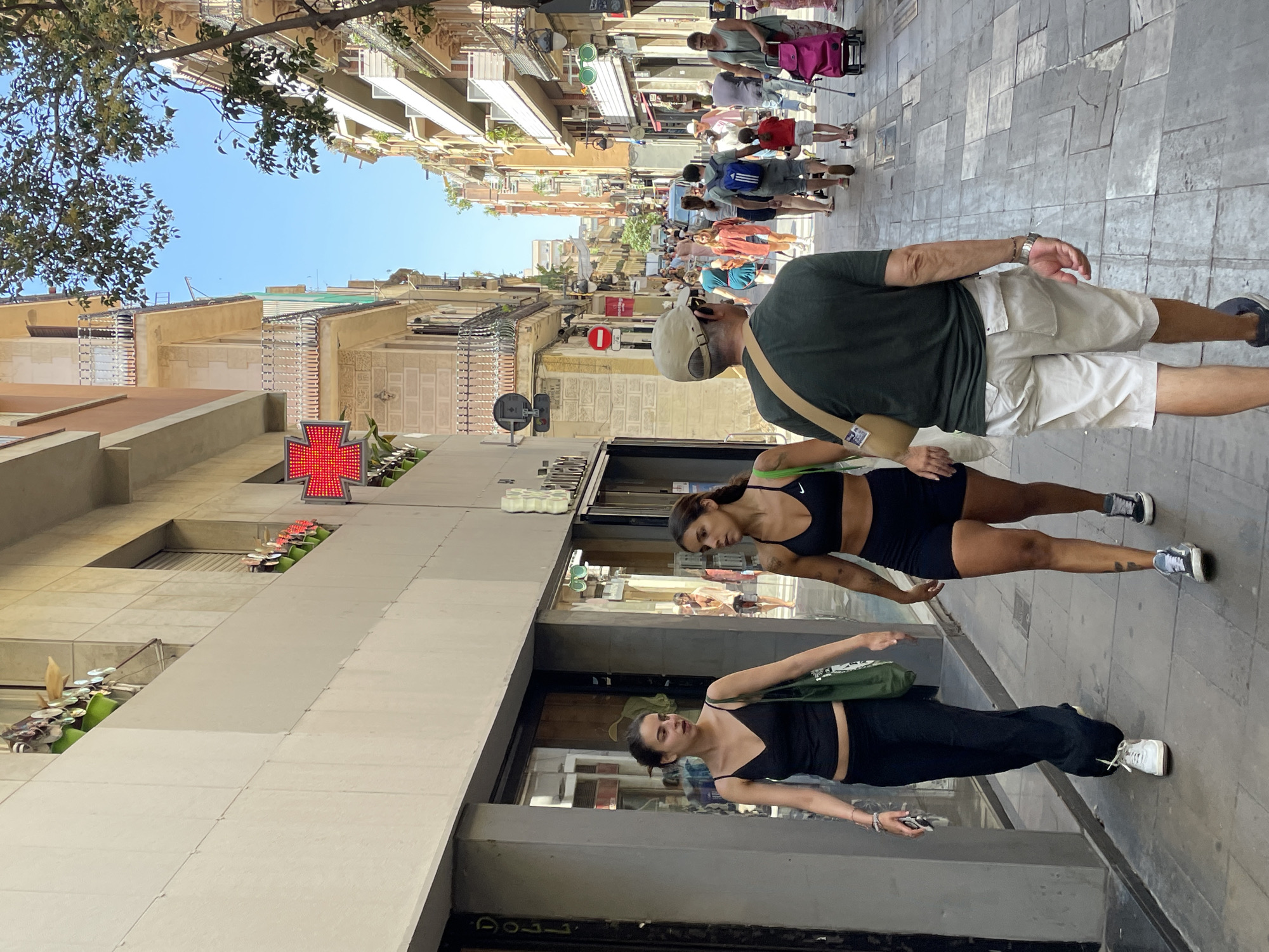 This is a medium shot, eye-level, outdoor photograph taken on a sunny day in Barcelona, Spain, likely on a pedestrian street lined with multi-story buildings. The scene depicts several people walking along a cobblestone sidewalk. In the foreground, a man wearing a light-colored baseball cap, a dark green t-shirt, and light-colored shorts is walking away from the camera. To his left, two women are walking towards the camera. The woman on the left wears a black tank top and black pants. The woman on the right wears a black sports bra and black shorts. Behind them, other pedestrians can be seen walking in both directions, some pushing shopping carts or strollers. The buildings on either side of the street have balconies and windows, with some greenery visible on the upper levels. A large red cross sign is mounted on one of the buildings. The overall impression is of a busy urban environment on a pleasant day.
