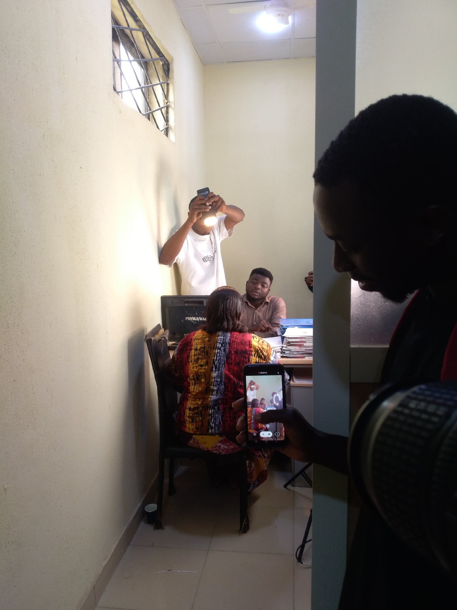 The image depicts an indoor scene in Jos, Nigeria, likely a production set for filming or photography during daytime. In the immediate foreground on the right, a person is partially visible in silhouette, holding a smartphone with their left hand to record the scene. The phone's screen displays a live video feed of the ongoing shoot. Their right hand holds part of a larger camera or lens.
In the midground, several individuals are engaged in the production. A man in a white t-shirt, possibly bearing "Holy S" text, stands against a light-colored wall, holding a bright flashlight or LED panel to illuminate the subjects. Below him, a person, seen from the back, is seated on a chair, wearing a vibrant, multi-colored patterned garment. They face a computer monitor on a desk, which shows the text "PSP/BLD/MAC". Adjacent to them, another man in a light brown shirt is seated at the desk, looking towards the back-turned person. The desk is organized with stacks of papers or binders.
The setting features light-colored, cream-like walls and a tiled floor. A window with dark security bars on the upper left allows natural light into the room. A ceiling fan with an integrated light fixture is visible towards the top right. A small, dark cup is on the floor near the wall. The arrangement suggests a collaborative effort to capture a scene, potentially an office interaction or an interview, with dedicated lighting and multiple recording devices.