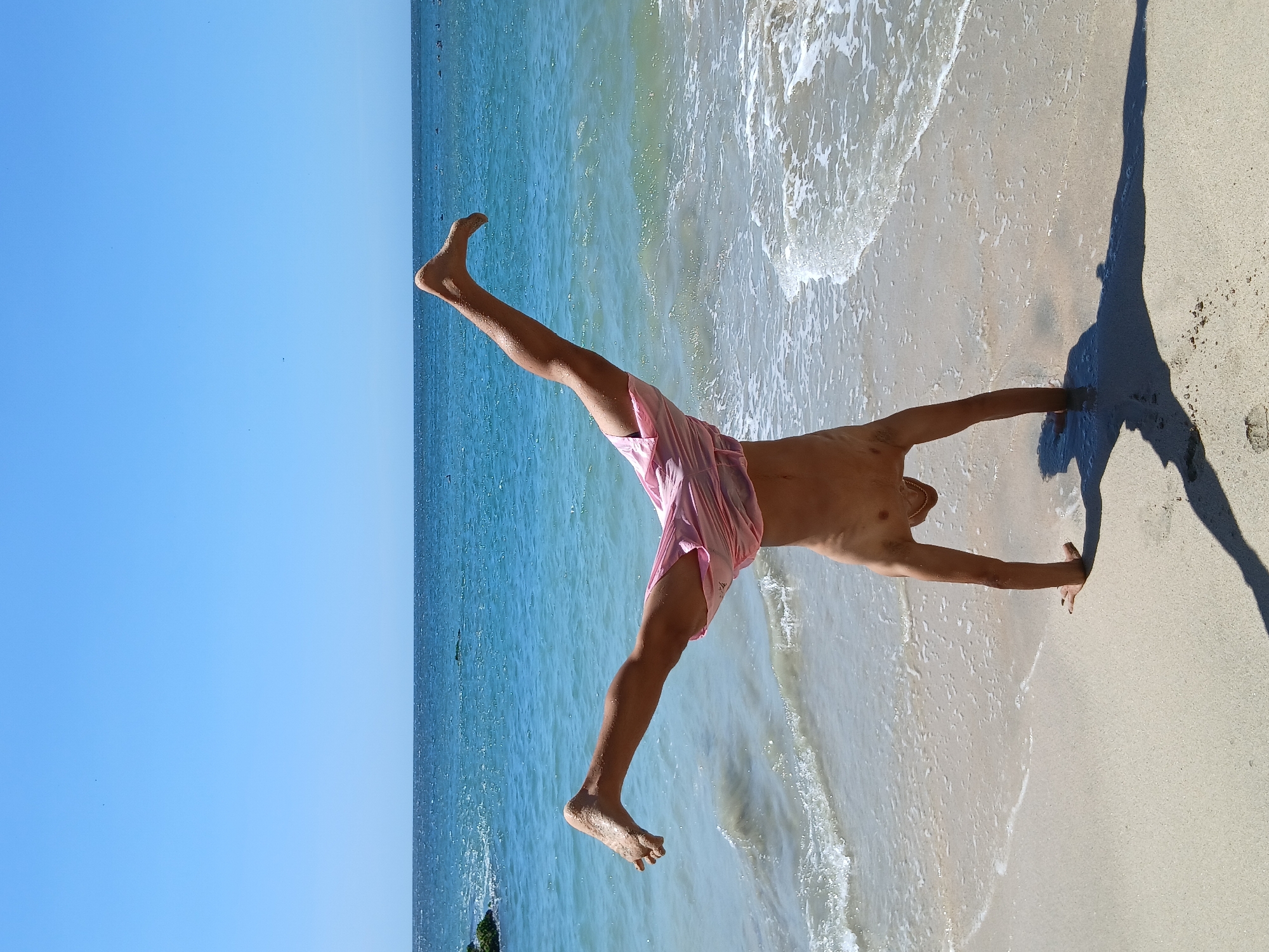 A person is captured in an athletic, inverted pose on a sunny beach, with the ocean and a clear blue sky forming the background. The individual, a shirtless male wearing pink shorts, is balancing on his right hand with his left arm extended forward, both hands pressed into the wet sand. His legs are spread wide apart, forming an inverted "V" shape, suggesting a handstand or a similar acrobatic straddle pose.
The setting is a vibrant beach scene under bright daylight, indicated by the strong sunlight and the distinct, elongated shadow of the person cast on the light-colored sand to the right. The sky is a uniform, clear blue, meeting the calm, turquoise waters of the ocean. Gentle waves are visible, breaking onto the shore and creating white foam patterns near where the person is performing. A small, dark mass, possibly rocks or distant land, can be seen in the water towards the left. The scene evokes a sense of activity and enjoyment in a coastal environment, consistent with a location like Pedro González, Venezuela. There is no visible text in the image.