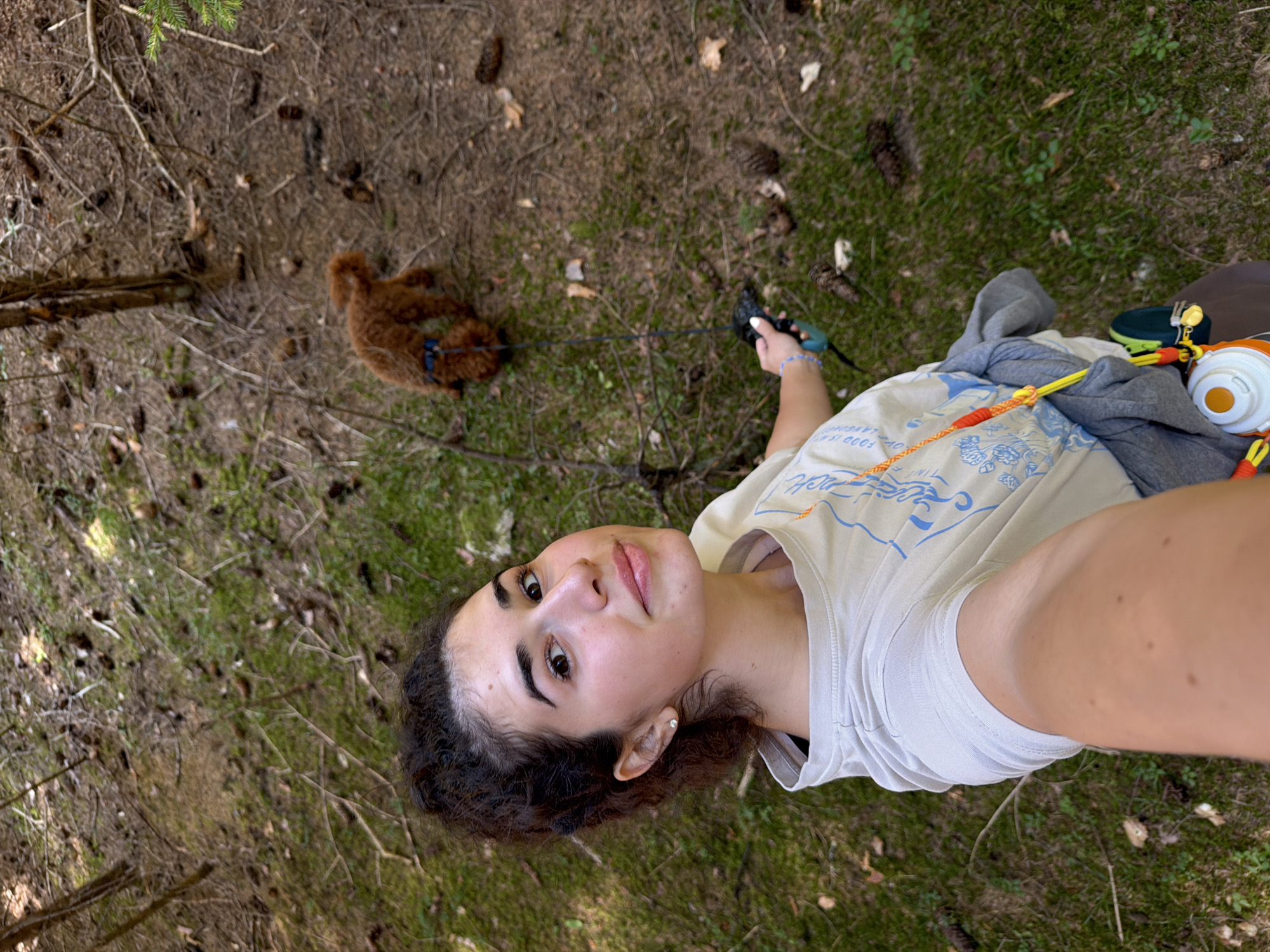 In Sarnitsa, Bulgaria, a young woman with dark curly hair tied back in a bun takes a selfie in a wooded area. She is wearing a light-colored t-shirt with a blue graphic design and a grey jacket tied around her waist. Her left arm is extended, holding a dog leash attached to a small, fluffy brown dog walking ahead of her. The ground is covered in moss, pine needles, and pine cones, indicating a forest environment. A water bottle is attached to a strap around her waist.