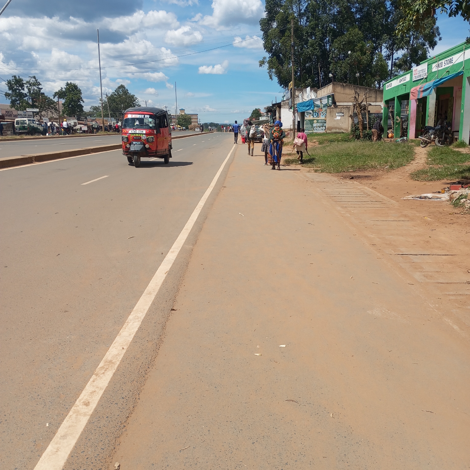 The image shows a street scene in Sheywe ward, Kenya. A red tuk-tuk is traveling on the left side of a wide paved road with a white line marking the edge. On the right side of the road, several people are walking, some carrying goods. Alongside the road are several small businesses, including a "FOOD STORE" with green and white paint and the word "AINYOZI" on its façade, as well as an electrical shop with a covered awning. A motorcycle is parked outside the "FOOD STORE". Trees are visible in the background. The sky is partly cloudy. Other vehicles and people are visible further down the road. A sign reading "HOSPITAL" and "MOKYS HIGHWAY SHOP CLINIC LANG" is seen on a building in the distance. A poster related to FIFA can be seen on the side of a building.