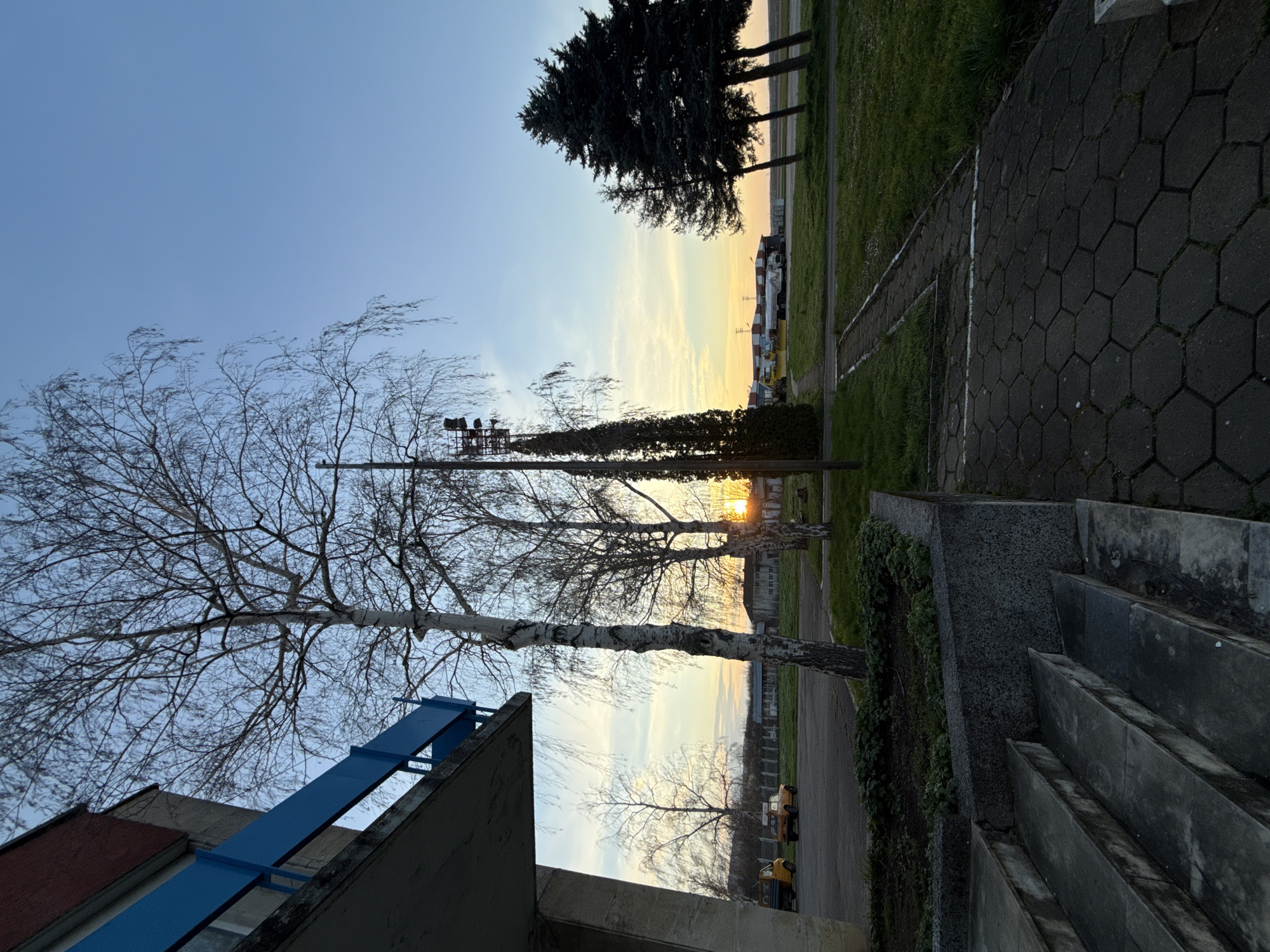 The image shows a cityscape scene, possibly taken in Gorna Oryahovitsa, Bulgaria. The photograph is rotated and presents a mix of natural and built environments. In the foreground, there's a concrete structure with blue railings, possibly a part of a building. Bare-branched trees are prominent against the sky. The background reveals a gradient sky suggesting sunrise or sunset, and a row of buildings and machinery. A paved area with hexagonal patterns and a grassy area with trees are visible further back. A concrete staircase leads towards the background.