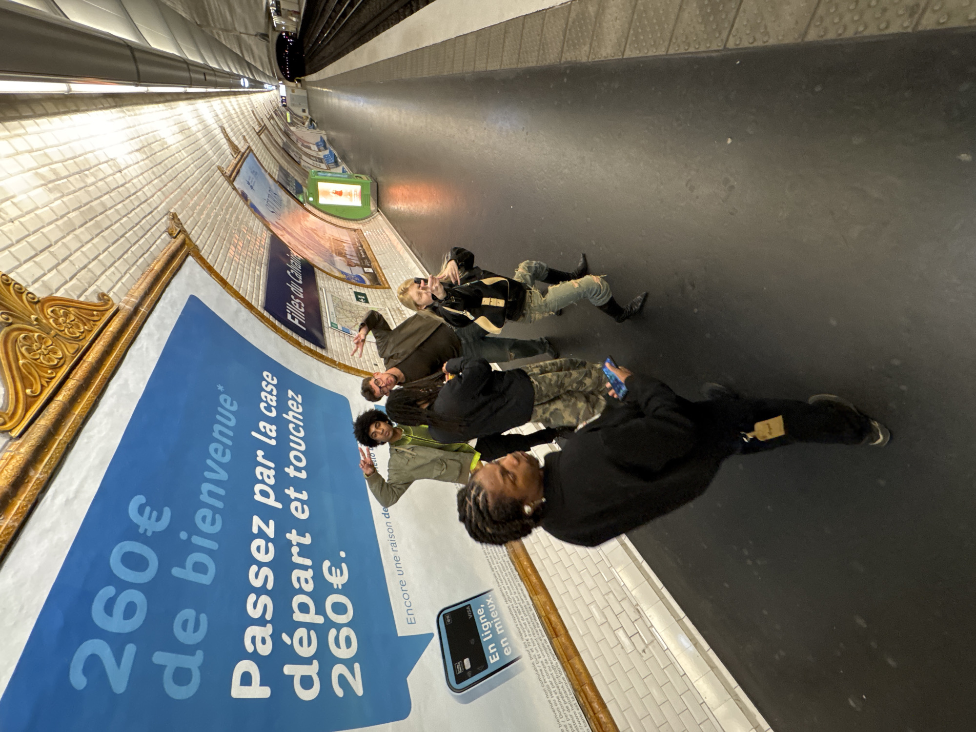 The image captures a group of five people lying on the floor of a Parisian metro station. They are positioned near an advertisement on the tiled wall which announces "260€ de bienvenue, Passez par la case départ et touchez 260€," advertising a promotion from Hello Bank Visa. The station's wall is decorated with vintage advertisements in ornate gold frames. The floor is dark and smooth. Overhead, the station's curving ceiling is visible, with a track for the metro. The station's name "Filles du Calvaire" is visible in one of the advertisements. The people are varied in age and dress and are facing the camera, some with their arms raised.