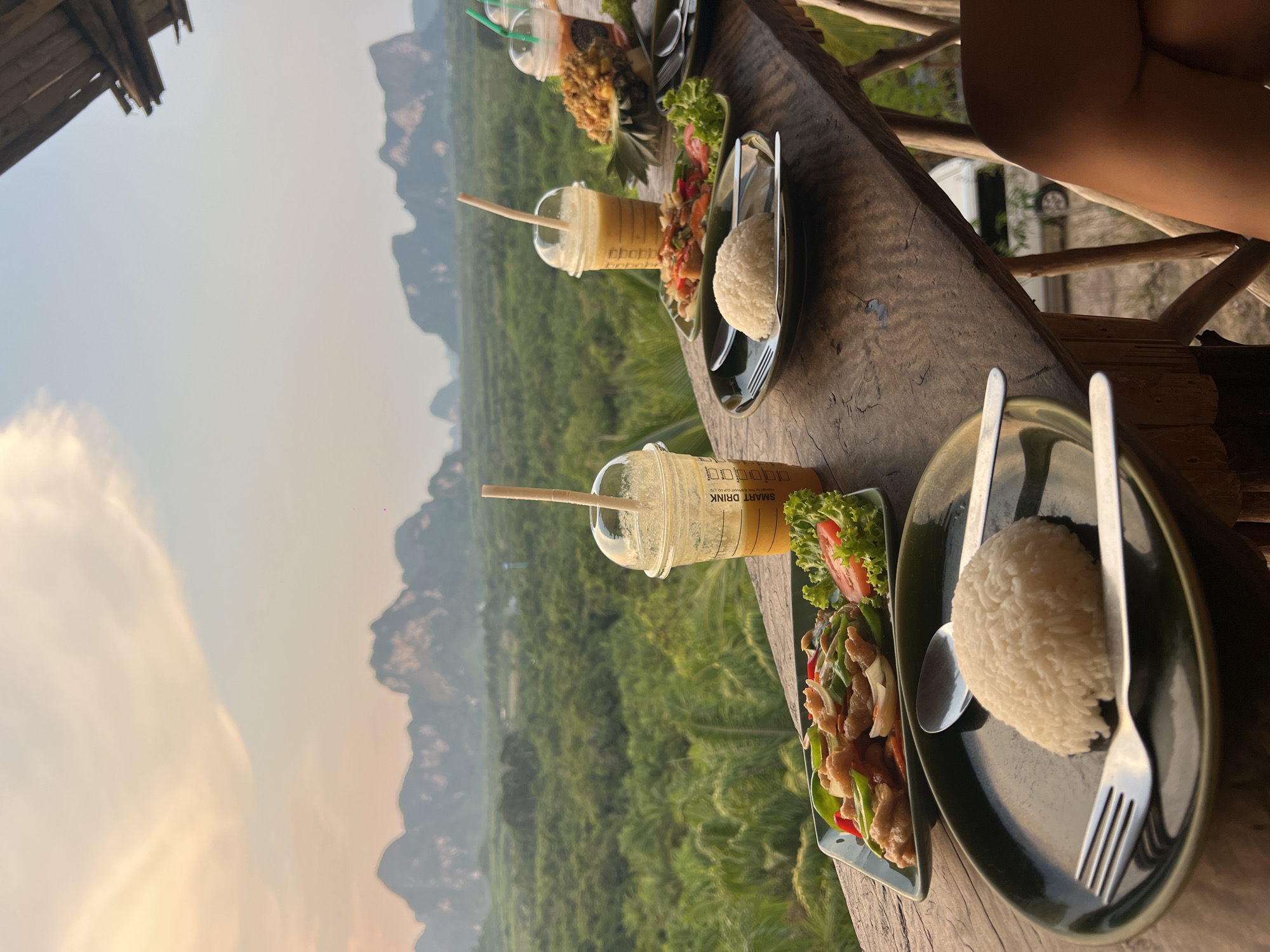 The image shows a table setup for a meal, with a backdrop of the Khao Thong landscape in Thailand. The table is made of dark wood, with the natural wood grain visible. Two plates are visible, each containing a portion of white rice and a side dish of meat stir-fried with vegetables, garnished with lettuce and sliced tomatoes. Next to each plate is a set of cutlery, including a fork and spoon. Adjacent to each plate is a tall, clear plastic cup filled with a yellow liquid, presumably a drink, and a brown straw. The cups have the words "SMART DRINK" printed on the side. The foreground is a balcony or raised platform, indicated by the weathered wooden railing. In the background, there is a lush, green valley with limestone karsts rising in the distance. The sky is a light, muted color, suggesting early morning or late afternoon lighting. A partial structure, possibly a roof, is visible in the upper left corner. A portion of a person's bare shoulder is visible in the top right of the image.