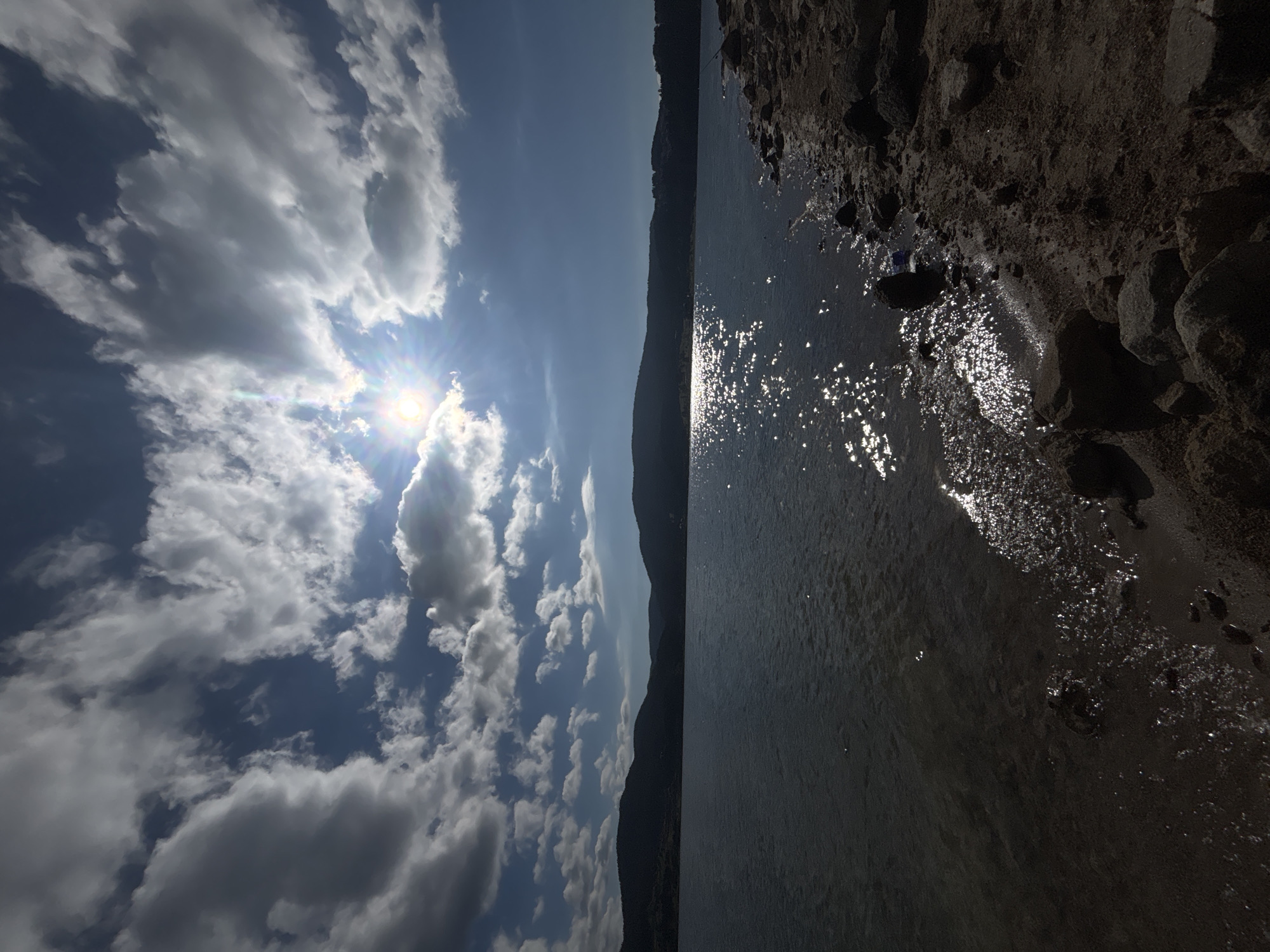 An ultra-wide-angle, low-angle shot captures a sunlit lake against a dramatic sky with cumulus clouds. The sun, positioned in the upper center, creates a dazzling reflection on the water. A dark, silhouetted mountain range stretches across the horizon, dividing the sky from the lake. The foreground is a rocky, sandy shore with scattered boulders. The overall scene suggests a natural landscape, possibly a reservoir or a large natural lake, under a dynamic sky.