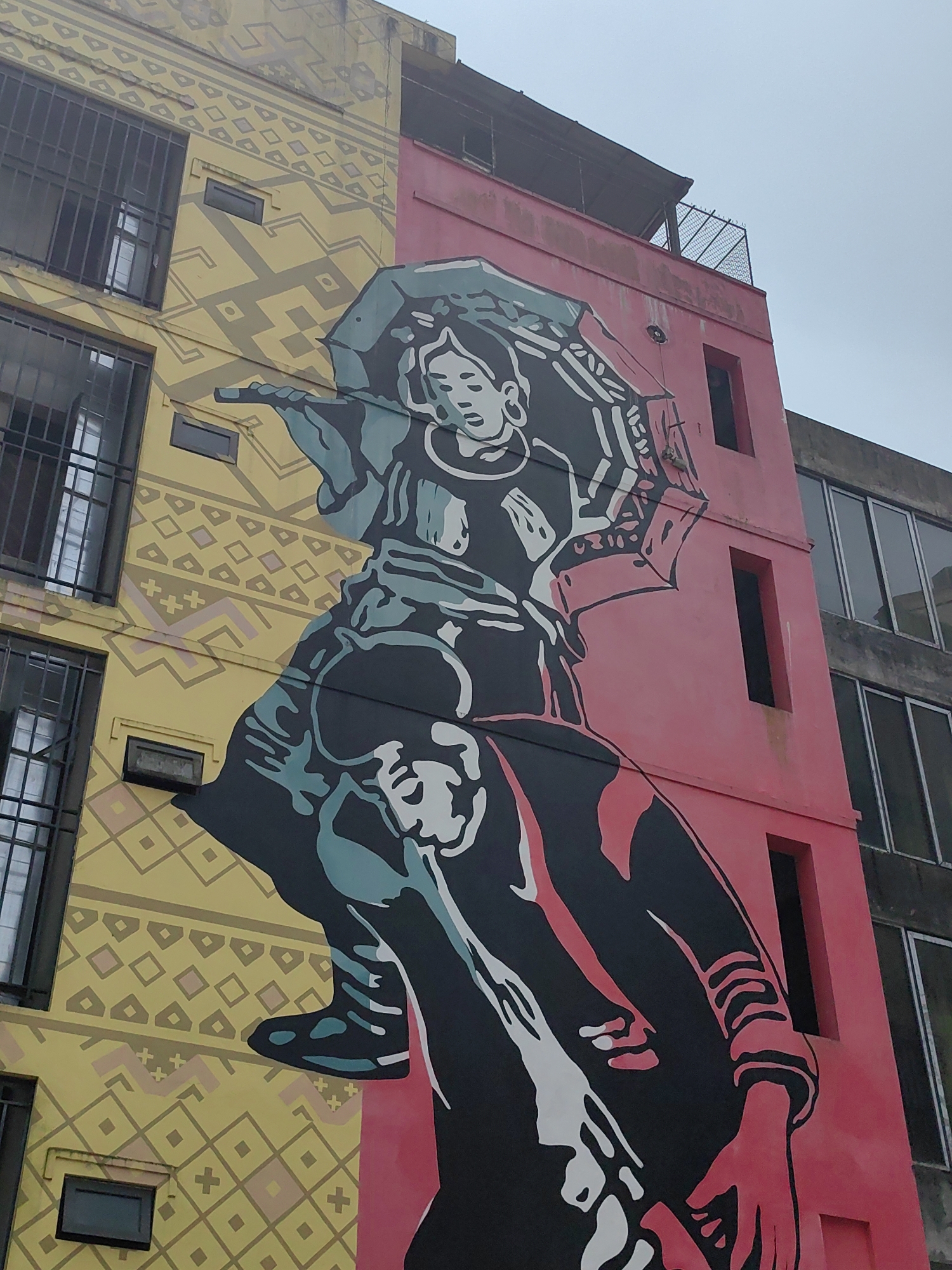 This image depicts a large mural painted on the side of a multi-story building in the city of Sa Pa, Vietnam, under an overcast sky. The building's facade is vertically divided into two distinct sections. The left section is yellow, patterned with intricate light brown geometric designs, and features multiple windows fitted with dark metal security bars. The right section is solid pink/red and contains several rectangular openings or recesses without visible glass or bars. An adjacent, unpainted concrete building with large windows is partially visible to the far right. The rooftop of the pink section includes a dark, sloped covering and a section of metal mesh fencing.
The mural itself spans both colored sections of the building, rendered in a graphic, high-contrast style using black, white, light blue-green, and a red-pink accent color matching the building's right side. The upper figure portrays a stylized woman, depicted from the waist up, wearing traditional attire. She holds an open, patterned umbrella with her right hand, while her left hand is also raised. Her foot is positioned above a second, lower figure who appears to be bent forward or carrying a load. The lower figure is predominantly black with red-pink accents on the arms and hands.