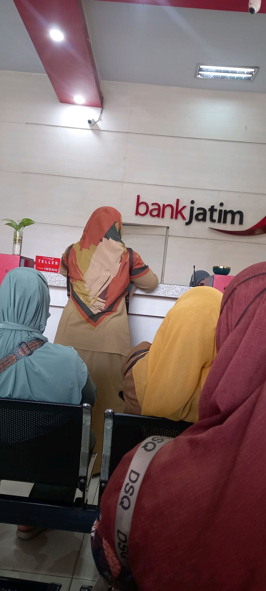 In a brightly lit interior, possibly a bank in Ambulu, Indonesia, several individuals are visible. The focal point is a counter area where a woman wearing a tan uniform and an orange and brown patterned hijab stands. In front of her, another person in a light blue hijab is partially visible, seated in a black chair. To the right, two other individuals, one in a yellow hijab and another in a maroon hijab, are also present. The maroon hijab features a distinct white band with the lettering "DSQ" repeated multiple times. Behind the counter, the "bankjatim" logo is prominently displayed in red and black. A small potted plant in a glass vase sits on the counter to the left, next to a sign that reads "TELLER INDAH." The overall environment appears clean and modern, with white walls and ceiling lights. The presence of surveillance cameras mounted on the ceiling suggests a focus on security within the establishment.