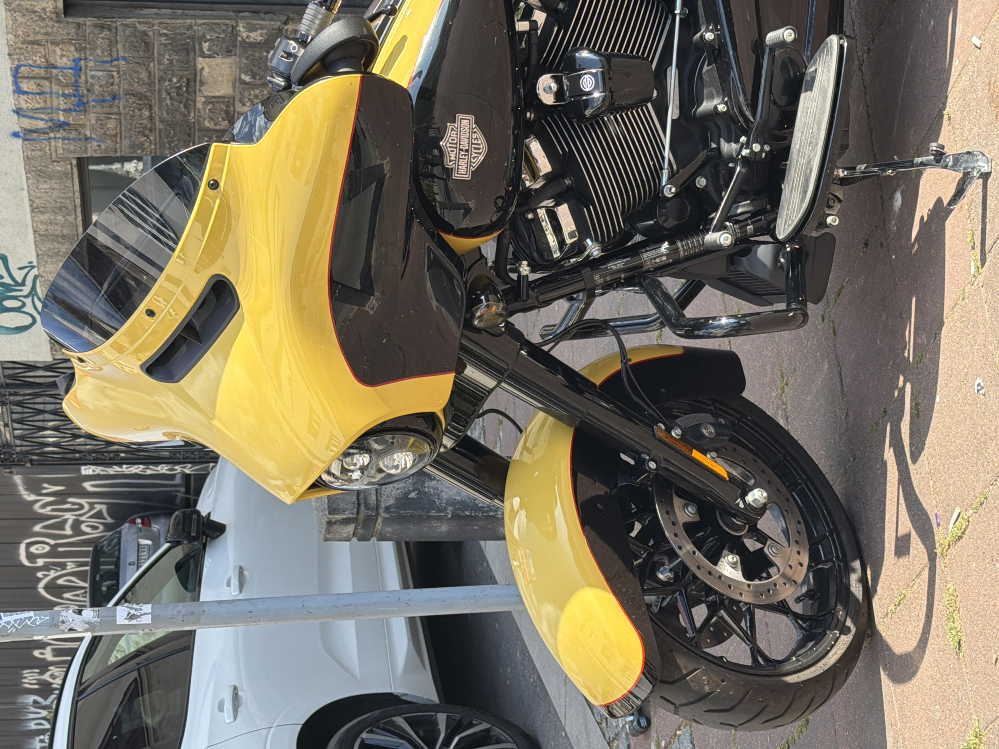 The image shows a close-up of a yellow and black Harley-Davidson motorcycle parked on a sidewalk. The foreground is dominated by the motorcycle's front end, including the yellow fairing, headlight, front fender, and black front tire with a visible disc brake. Parts of the engine are also visible. In the background, there is a white car parked next to the motorcycle, and behind the car, there's a building with graffiti-covered walls. The sidewalk is made of stone pavers. Based on available context, the location can be identified as Belgrade, Serbia.