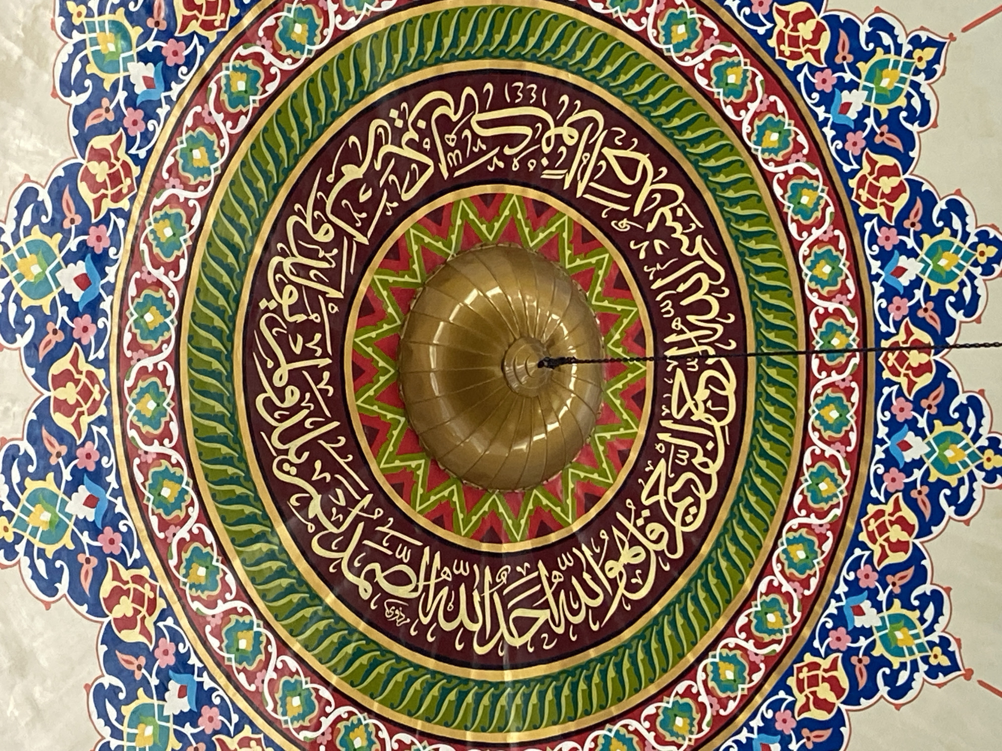 The image shows a close-up view of the ornate ceiling of the Baiturrahman Grand Mosque in Lhokseumawe, Indonesia. The ceiling is a circular design featuring intricate geometric patterns and Arabic calligraphy in gold lettering against a dark burgundy background. The calligraphy is surrounded by concentric bands of green, gold, and red with stylized floral motifs. At the center of the dome is a golden finial, suggesting the architectural style is consistent with Islamic religious buildings. The overall impression is one of rich detail and religious artistry.