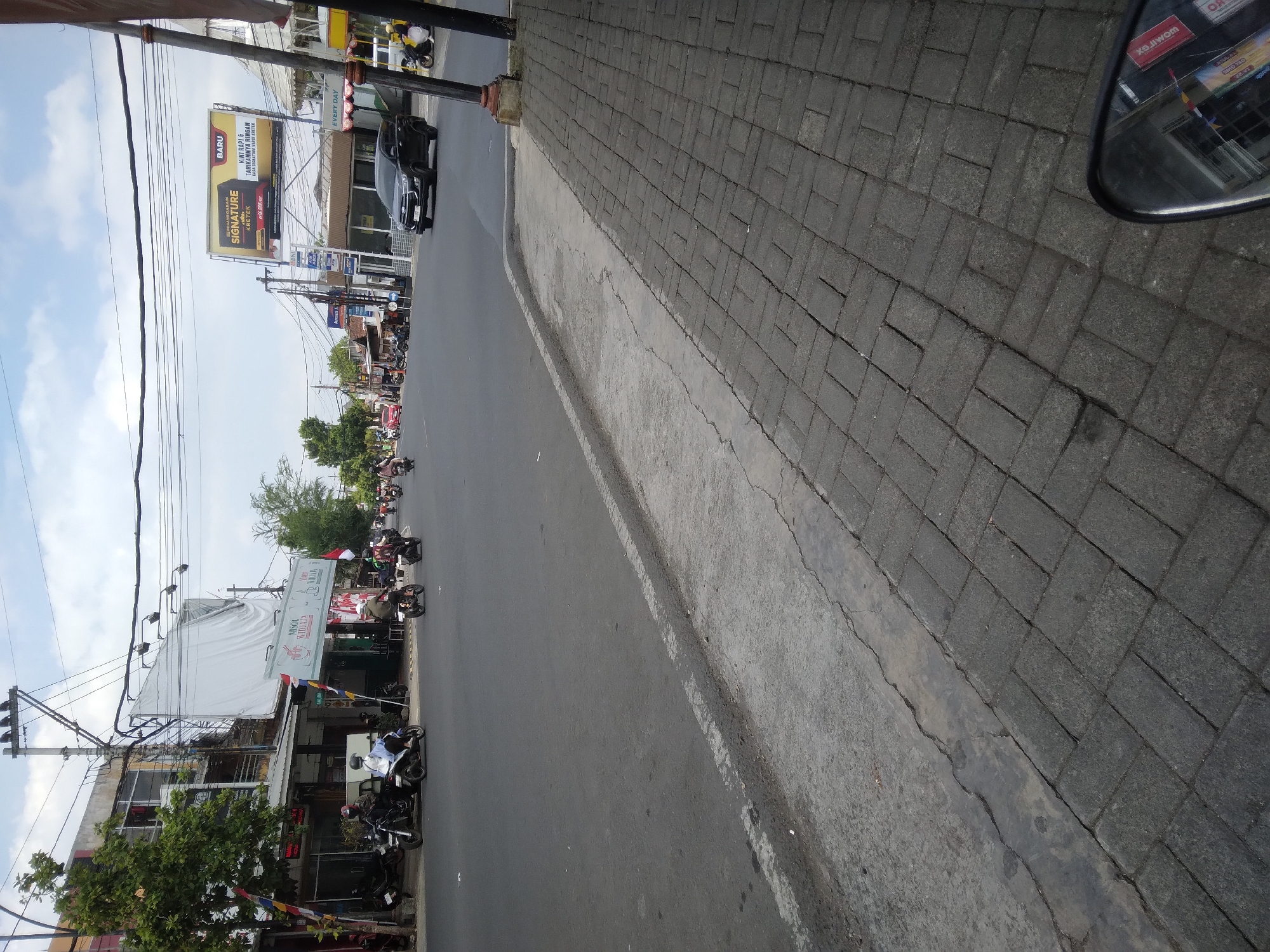 The image depicts a street scene in Berkoh, Indonesia. The street is paved with asphalt and lined with a brick sidewalk. Several motorcycles and a black car are visible on the road. Buildings with storefronts and signage line the left side of the street. A large billboard for "Signature Kretek" cigarettes is prominently displayed on the right side of the road. Overhead, a complex network of power lines crisscrosses the sky. The sky is partially cloudy. The overall scene suggests a typical urban environment with commercial activity and traffic.