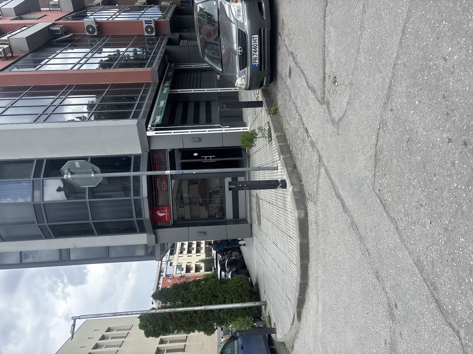 The image depicts a street scene in Plovdiv, Bulgaria, featuring a modern building with glass windows and a dark gray car parked in front. The license plate number of the car is PB7440HT. The building houses a business, possibly a restaurant or cafe, as indicated by signage. A street sign is visible near the building. On the opposite side of the building are additional buildings with greenery and parked cars. The pavement shows signs of wear and tear with cracks and patches. There are people near the entrance to the building. The sky is overcast.