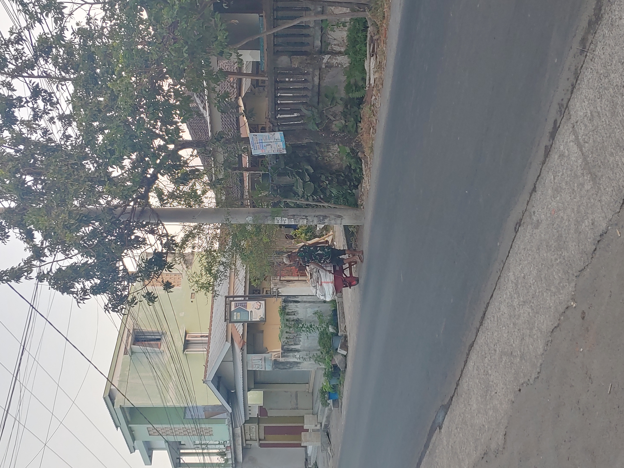 An elderly person, dressed in a camouflage-patterned shirt and dark pants, walks on a paved road in Kembang Kerang, Indonesia, pulling a red wheelbarrow filled with large sacks of what appears to be sand or other loose material. The person is walking away from a line of buildings on the left side of the road, which includes a light green, multi-story structure with balconies and windows, and a smaller, yellow building with a closed storefront. A tall utility pole stands near the wheelbarrow. The environment includes trees with dense foliage, a concrete wall with decorative elements, and a clear sky. The road is asphalt, with a concrete pedestrian path on the left.