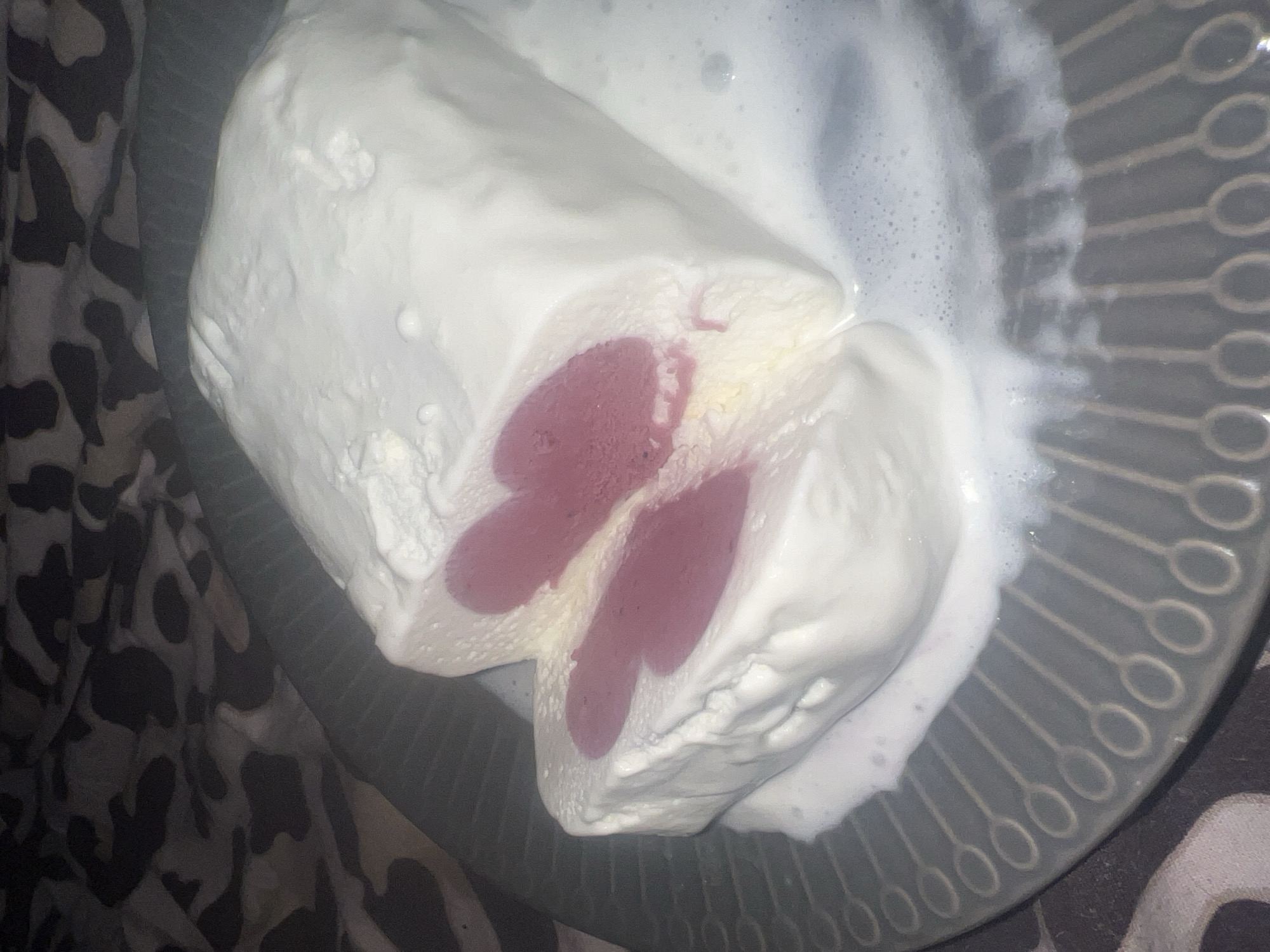An overhead shot captures a partially melted dessert on a dark grey plate, positioned against a patterned fabric. The dessert, likely a frozen confection such as ice cream or semifreddo, is a log-shaped treat with a creamy white exterior. It has been cut or broken open, revealing a vibrant pinkish-purple core, which appears to be composed of two distinct, elongated, heart-like or oval shapes. The white outer layer exhibits a soft, somewhat lumpy texture, and significant melting is evident as a frothy, white liquid pools around the dessert on the plate.
The plate itself is dark grey or black and features a decorative radial pattern of connected, raised oval shapes. Adjacent to the plate, and partially beneath it, is a fabric with an abstract, dark grey and black camouflage-like or organic pattern. The lighting is soft and indirect. No human subjects are visible, nor are there any discernible emotions or visible text. The scene depicts a sweet item in an indoor setting. This image was taken in Sinyavino, Russia.