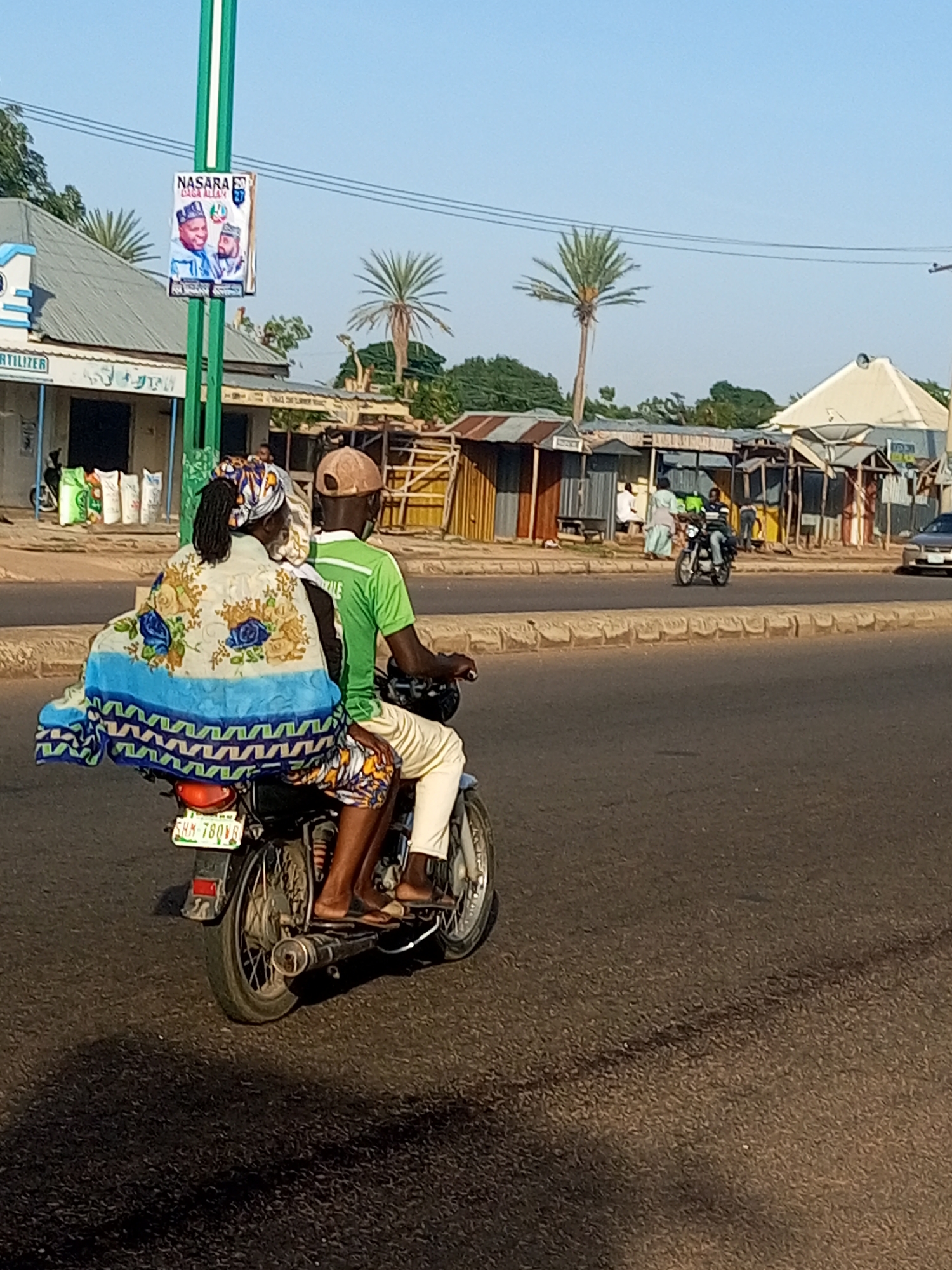 In Tempure, Nigeria, two individuals are riding on a motorcycle. The person in front, likely the driver, is wearing a green t-shirt and a light-colored cap. The person in the back is wearing a headscarf and a patterned wrap that drapes over the back of the motorcycle and the rider's legs. They are traveling on a paved road with shops and buildings visible on the left side of the road. A poster featuring two men's portraits is attached to a green pole on the left. Palm trees are visible in the background. The scene suggests a typical day in this Nigerian city.