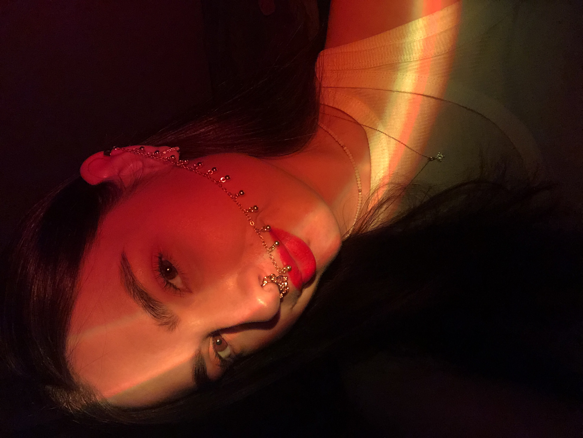 A medium shot captures a woman's face and partial torso, seemingly oriented at a tilted angle to the viewer. A chain adorns her left ear and the side of her nose. She is wearing lipstick. The lighting is a combination of red and yellow. She is wearing a white shirt. The rest of her body is obscured in darkness.
I do not have the information necessary to identify the location of Noviy in Russia from the image.