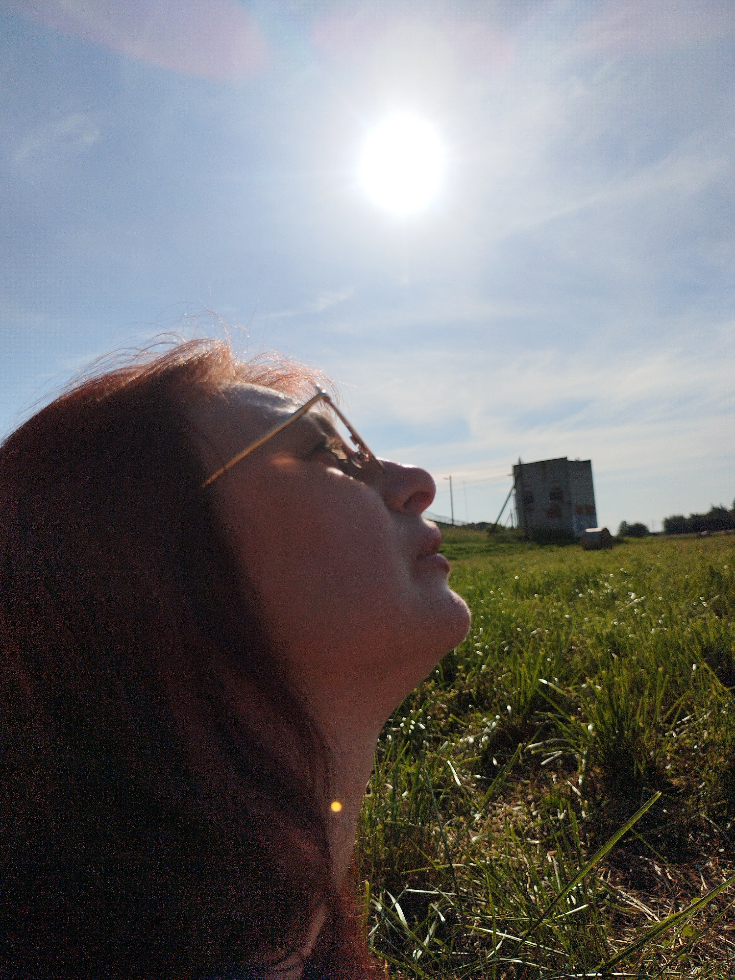 A low-angle, medium shot captures a woman with reddish-brown hair and eyeglasses looking up at a bright sun in a clear blue sky. She is positioned on the left side of the frame, with her face in profile. The sun is high in the sky and somewhat overexposed. In the background, a flat, grassy field stretches towards a slightly hazy horizon. A rectangular, two-story building with windows is visible in the middle distance. The grass is green and uneven, with some scattered white wildflowers. The overall lighting suggests a sunny day. The location is identified as Unknown, Unknown.