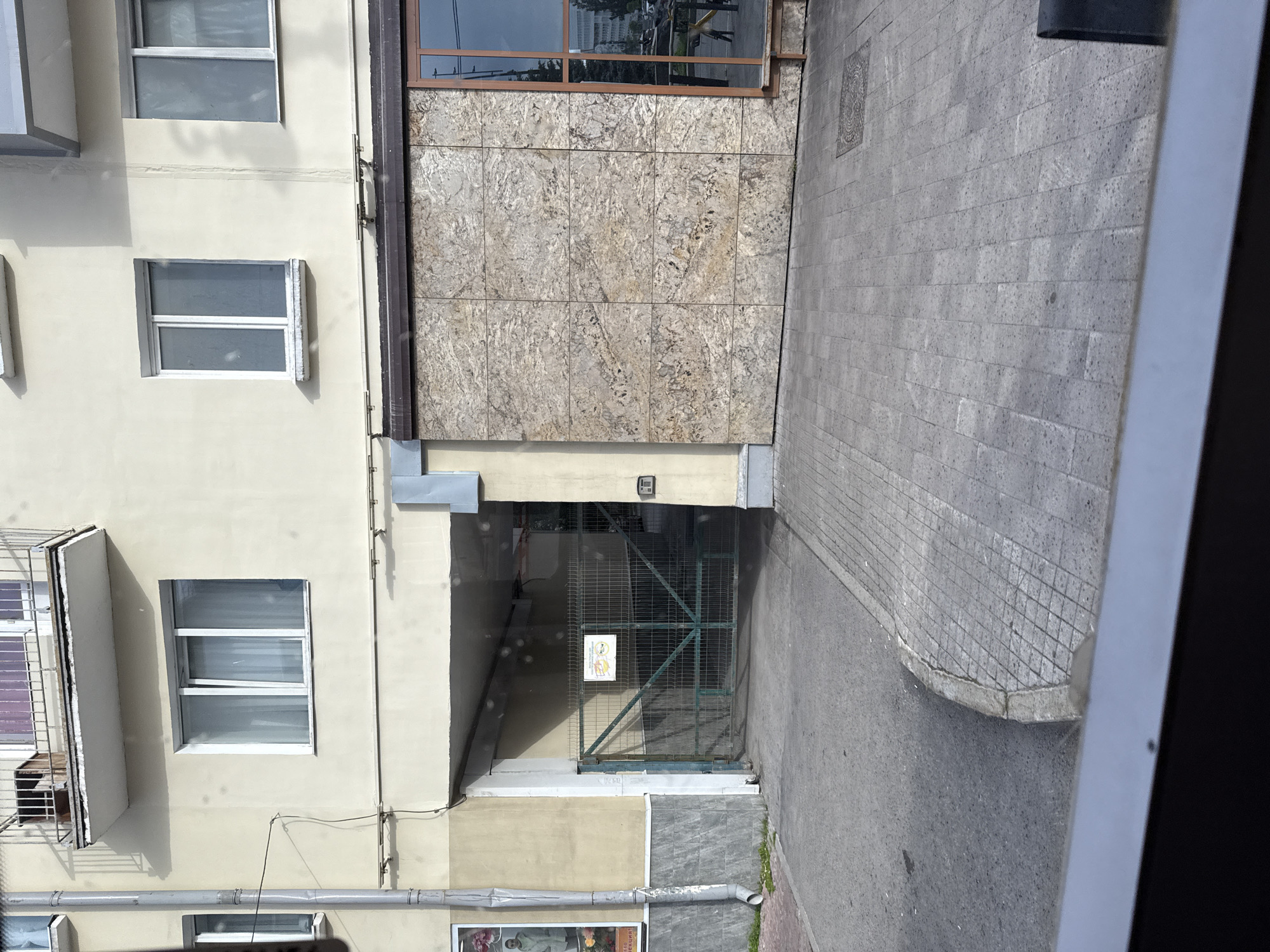The image depicts the facade of a building, likely an apartment complex, with a ground-level entrance and a paved walkway. The building has a pale yellow exterior with several windows on the upper floors. A balcony with a railing is visible above one of the windows.
The ground level features a covered entryway with a green metal gate. A sign with some text and images is attached to the gate. To the right of the entryway, a wall is covered with large, light-colored stone-like panels. A large window with a dark frame is situated next to this wall, reflecting the environment outside.
The walkway in front of the building is paved with rectangular stones. A curved concrete curb separates the walkway from what appears to be a road or parking area.
The overall scene suggests an urban environment. Without further context, it is impossible to definitively determine the exact location within the city of Perm, Russia. However, the architectural style and the presence of the metal gate are consistent with residential buildings found in Russian cities.