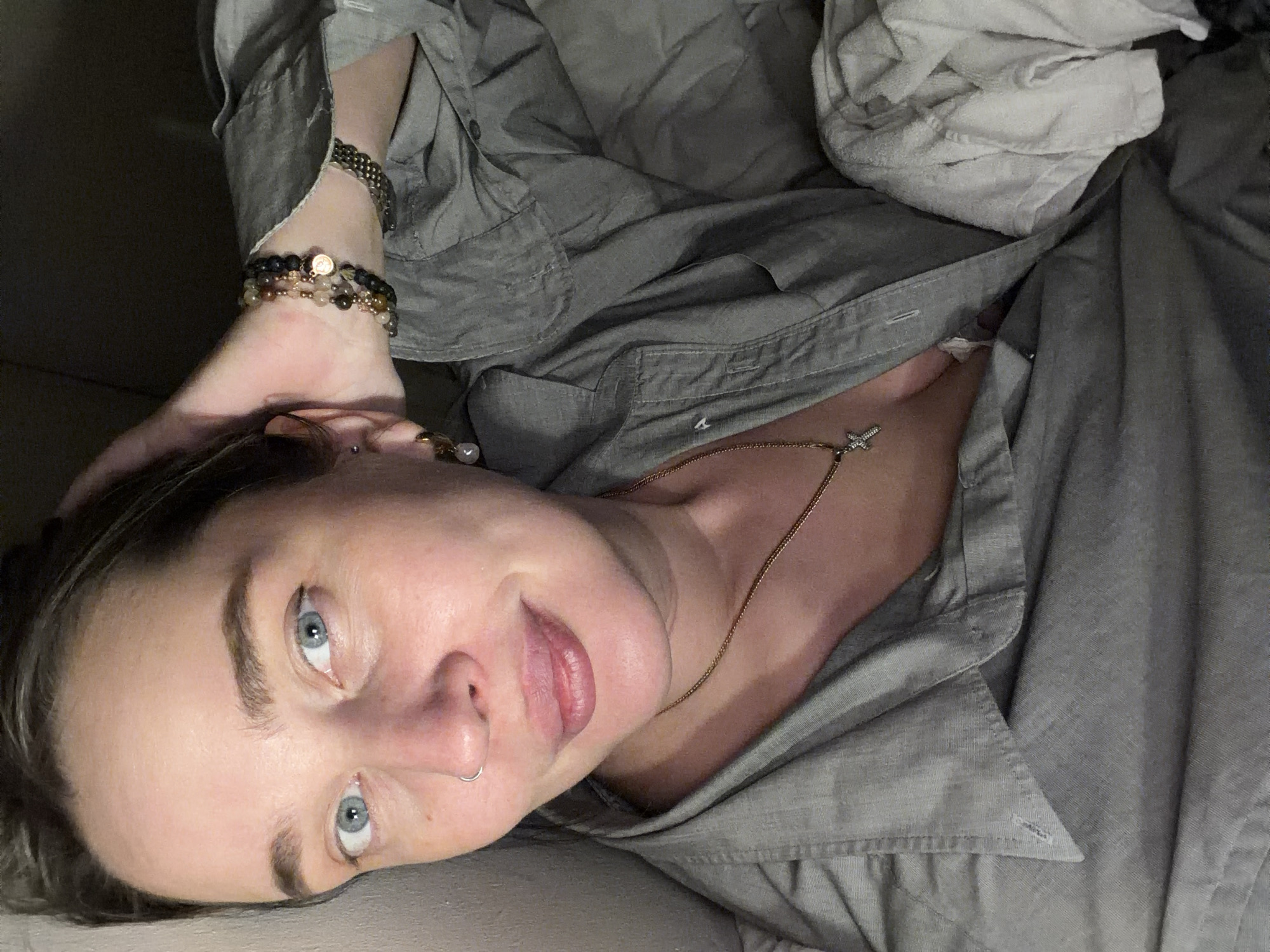 A person is lying down, facing the camera. They have fair skin, blue eyes, and brown hair. They are wearing a gray button-down shirt and a gold necklace with a charm. They have bracelets on their left wrist and a nose ring. Part of a white fabric or towel is visible near the upper right corner. The setting appears to be indoors. No information relates to Moscow.