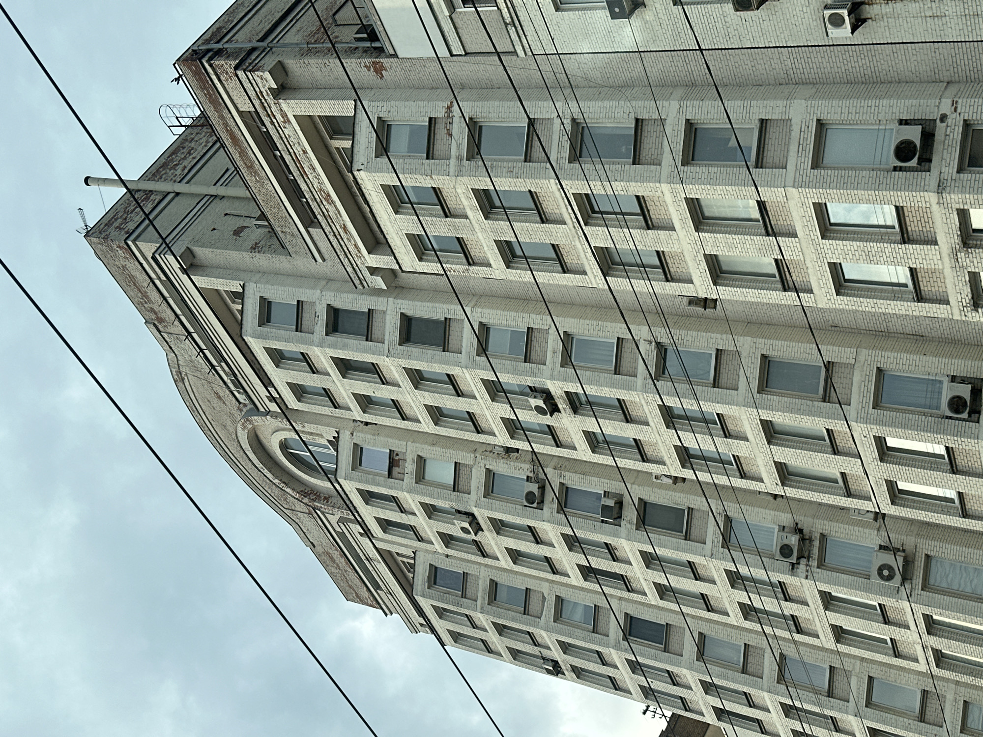 The image shows a tall, light-colored brick building, possibly located in Kyiv, Ukraine. The building has numerous windows, many of which have air conditioning units installed below them. Several power lines run diagonally across the front of the building. The sky is overcast. The building has a rounded corner and architectural details such as ledges and protrusions. There is a cylindrical structure on the top of the building.