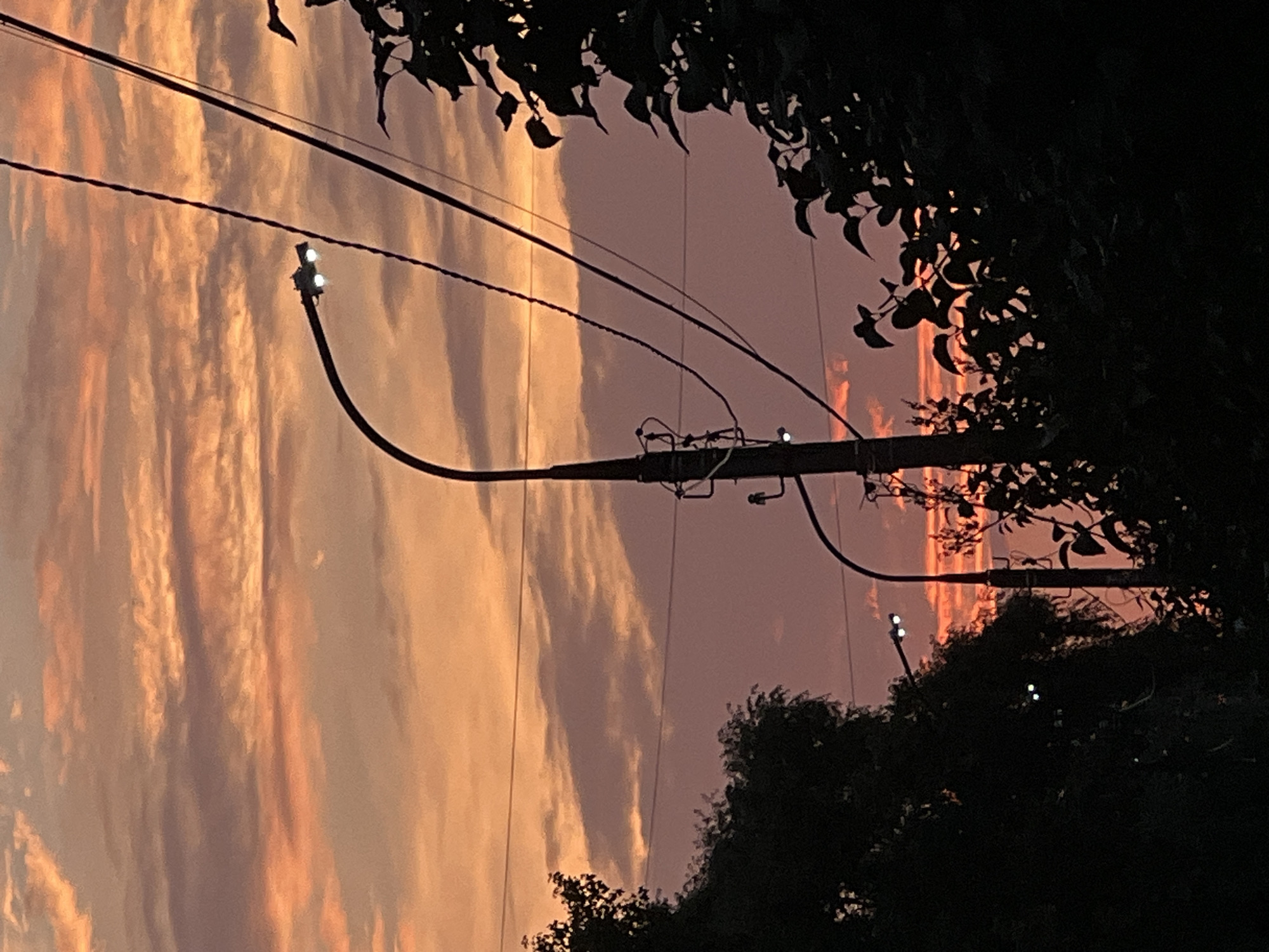 A street lamp with two illuminated bulbs is visible on the right side of the image, silhouetted against a twilight sky. The sky is filled with clouds displaying hues of orange, pink, and purple. Power lines are strung across the scene, and the dark silhouettes of trees frame the lower portion of the image. A second street lamp is partially visible on the lower left, also illuminated. The setting appears to be an urban or suburban area in Chaikovsky, Russia, given the presence of street lights and power infrastructure against a natural sky.
