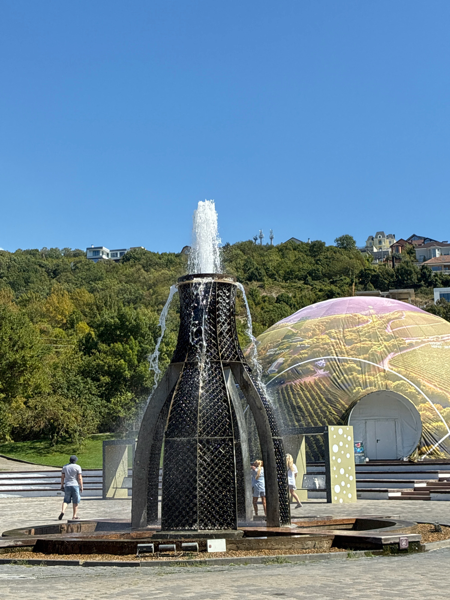 In Abrau-Dyurso, Russia, a large fountain shaped like a wine bottle is active on a sunny day, with water spraying upwards and cascading down its sides. In the background, a geodesic dome structure displays a vibrant image of vineyards. The dome is situated on a grassy hillside dotted with trees and several houses. To the left of the fountain, a man in a striped shirt and shorts walks away from the camera, while two women stand near the fountain. The ground around the fountain is paved with cobblestones.