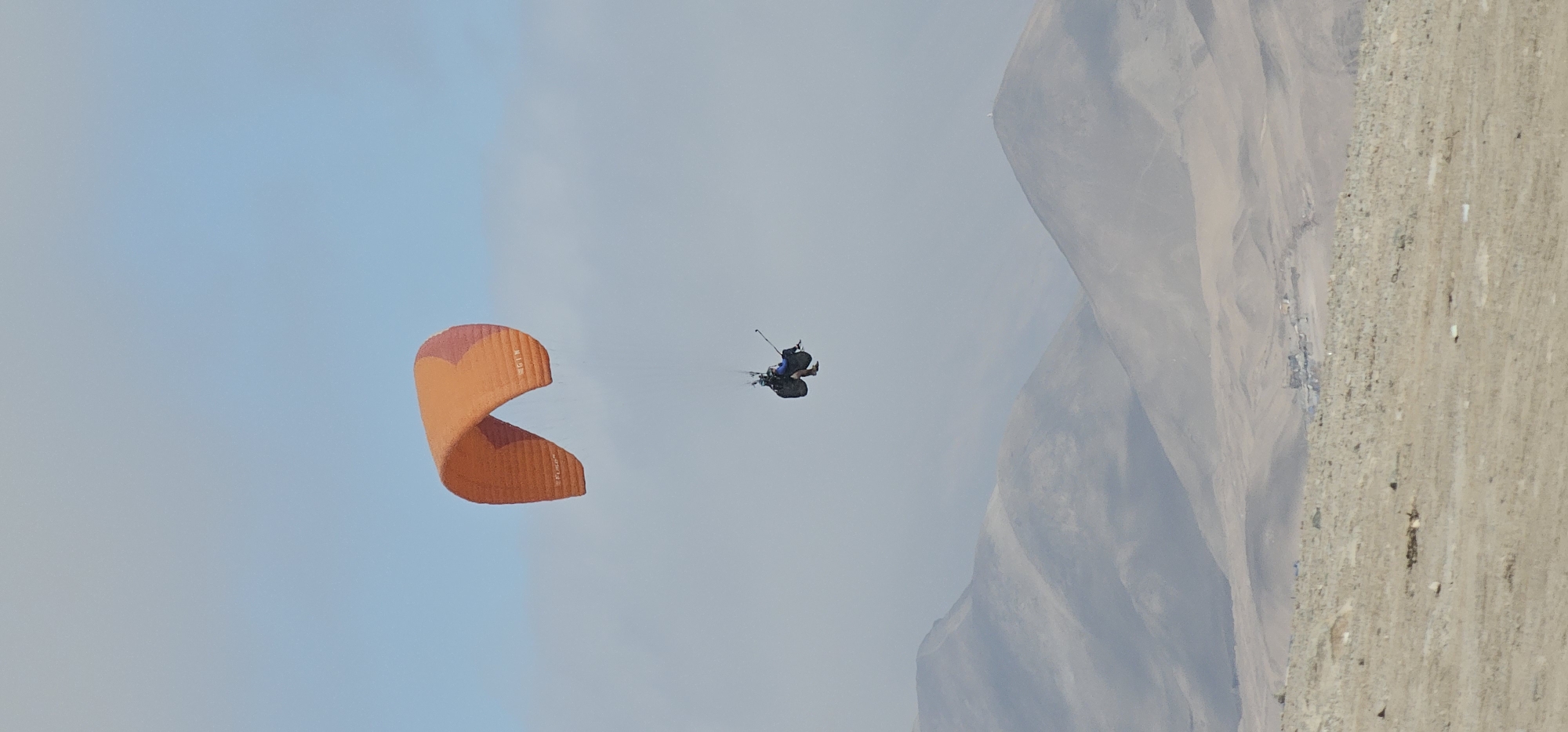 An aerial view shows a tandem paragliding flight occurring near Alto Hospicio, Chile. A paraglider with an orange canopy is in the air. Two people are visible strapped into the harness below the canopy. The environment includes a mountainous backdrop and clear blue sky. Some flat scrub and structures are visible on the ground at the foot of the mountains.