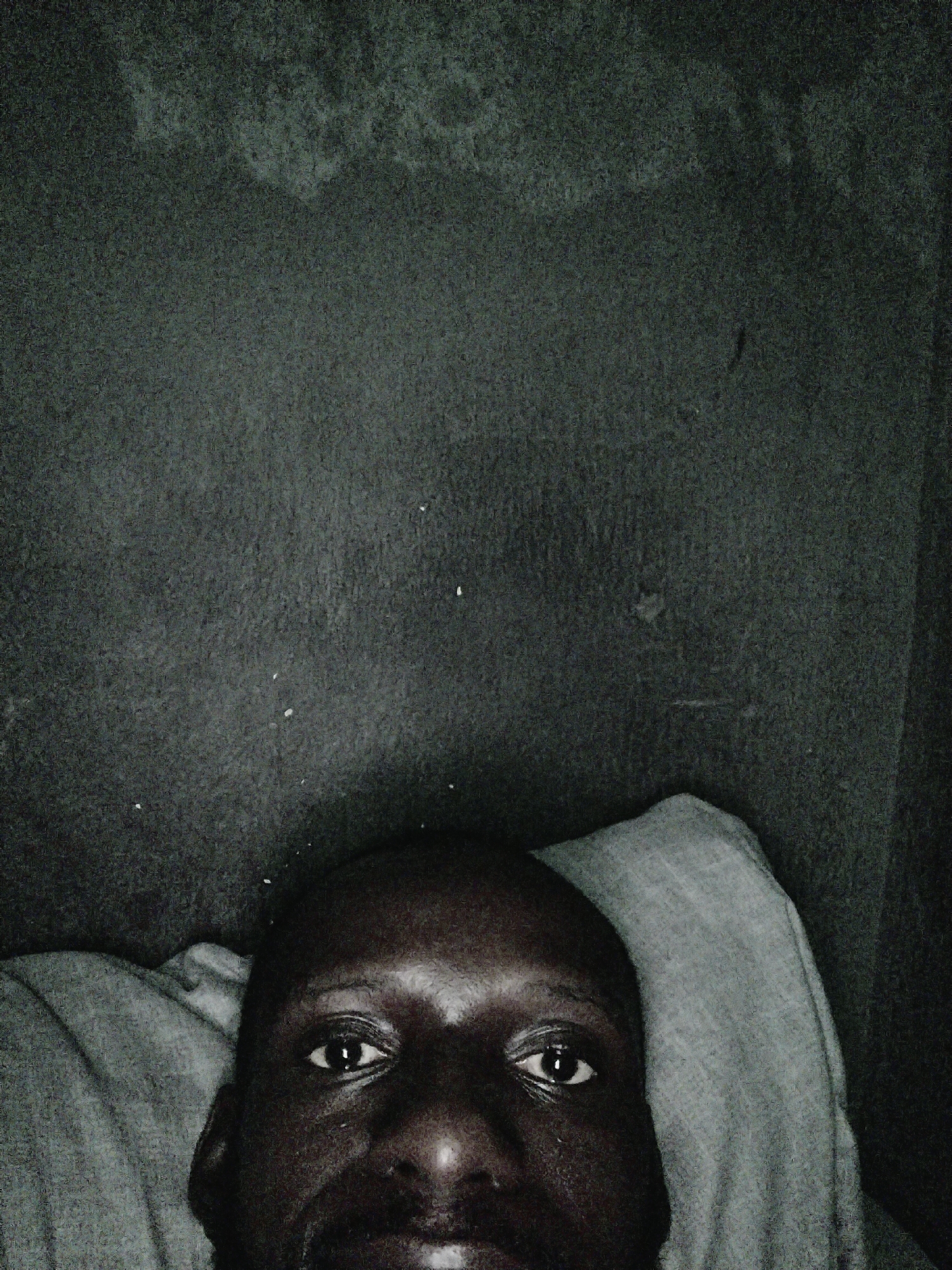 A close-up, low-angle, dimly lit, grayscale photograph depicts a Black male lying in bed. Only the upper portion of his head and face, from the forehead to the mouth, are visible. He is resting his head on a light-colored pillow. Above him is a dark wall with minor surface imperfections and a few scattered white specks. The location in the city of Uyo, Nigeria is undetermined.