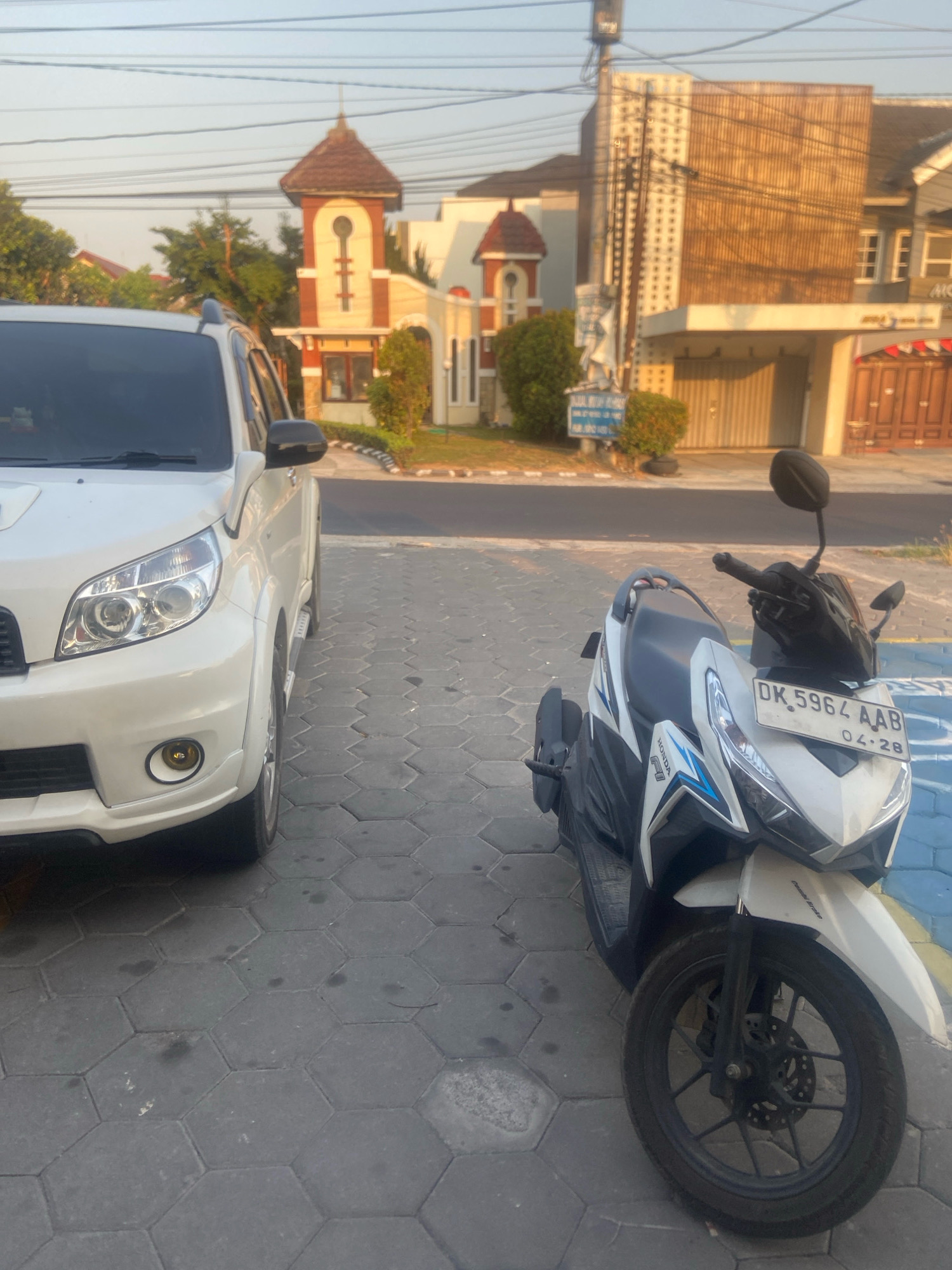 In Sleman, Indonesia, a white SUV is parked on a hexagonal paver driveway next to a white and blue scooter. The scooter has a license plate reading "DK 5964 AAB" and "04-28". Behind the vehicles, a two-story building with a modern facade and some architectural accents is visible. The building features a tan and brown exterior with several windows. A street with a road and sidewalk runs in front of the building. Trees and foliage are scattered throughout the scene, and overhead power lines are present. The sky is a clear, pale blue, indicating it is daytime. The overall scene suggests a residential or commercial area in the city.