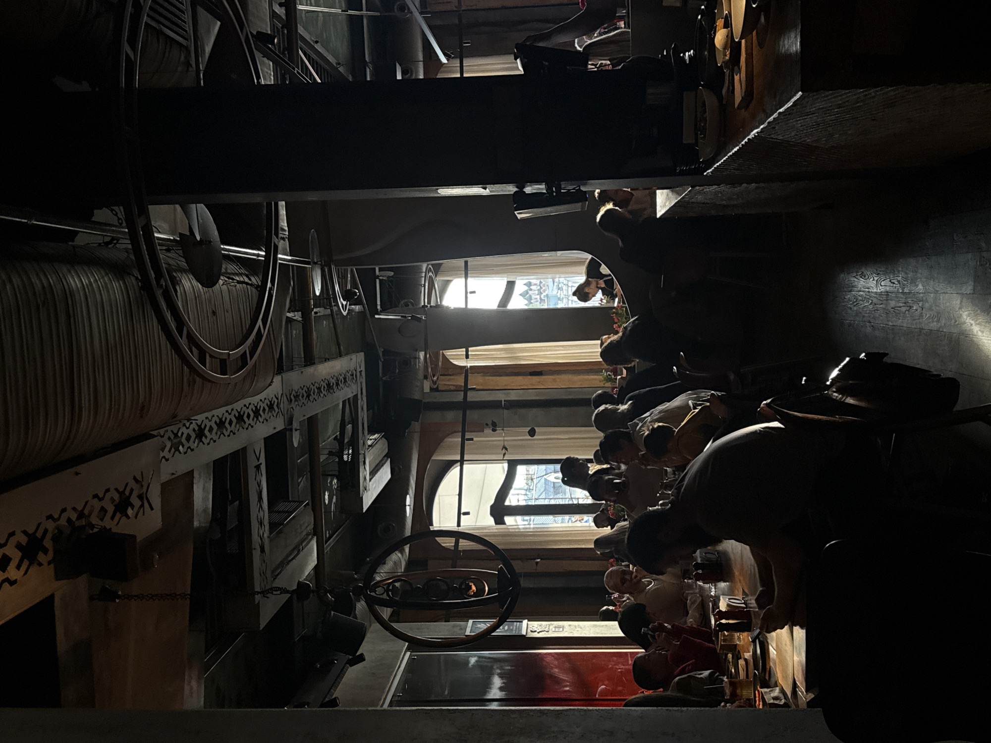 The image depicts a scene inside a restaurant in Moscow, Russia. The perspective is from a high angle, looking down into the establishment. The restaurant has a unique, possibly steampunk-inspired interior design, featuring large metal pipes, decorative patterns, and industrial-looking equipment. The lighting is dim, with light coming in from windows towards the back of the restaurant. Numerous people are seated at tables, seemingly engaged in conversations and dining. The seating arrangement suggests a crowded or popular venue. There are drinks, plates, and other dining accoutrements on the tables.