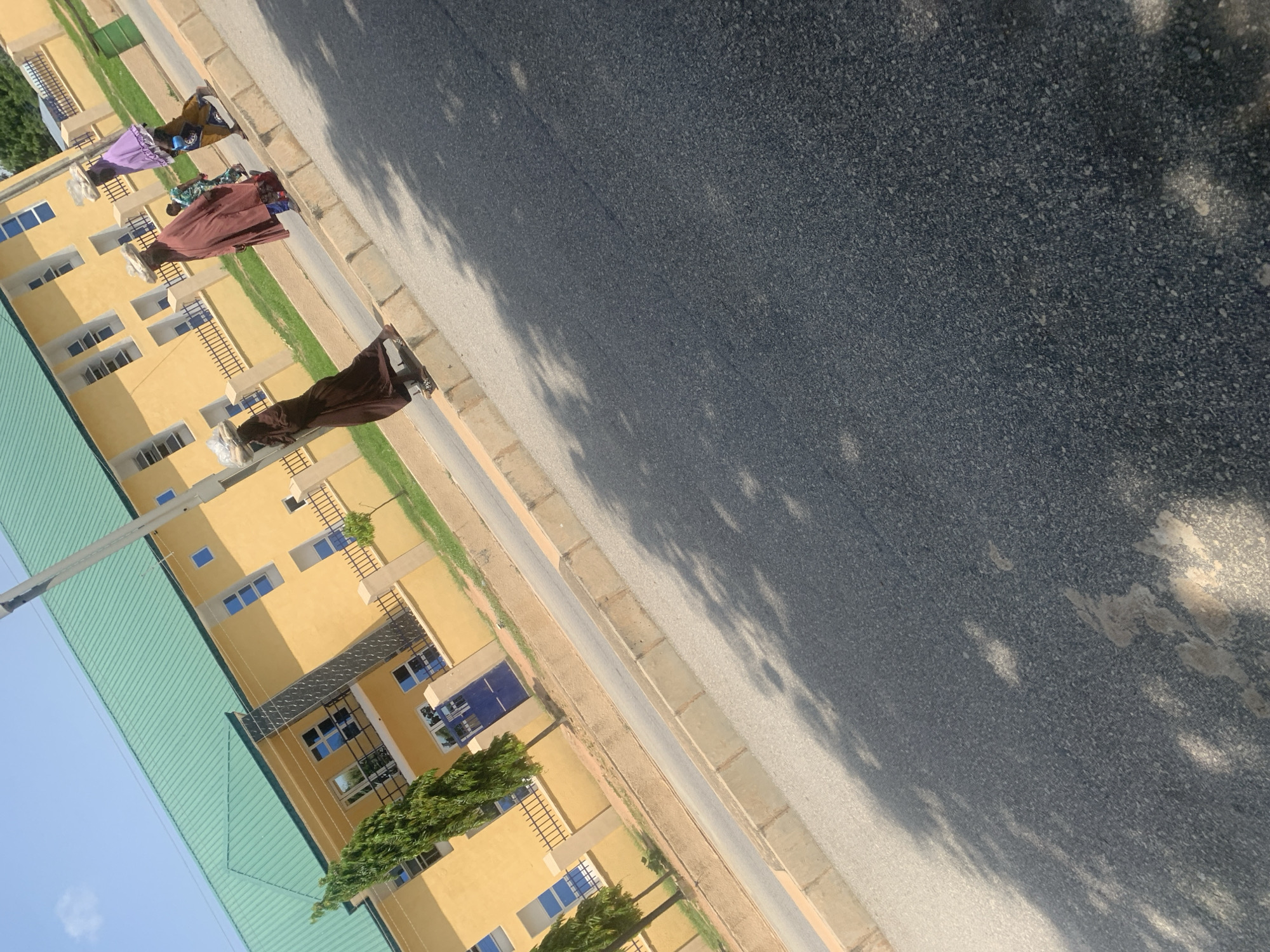 In Jalingo, Nigeria, on a sunny day, three individuals walk along a paved sidewalk adjacent to a yellow building with a green roof. The building has multiple windows and a dark blue gate. Two of the individuals, dressed in long, loose garments, carry large woven baskets filled with goods on their heads. One person, wearing a brown robe, walks to the left, while another, in a purple headscarf and brown skirt, walks to the right. Between them, a person in a patterned green and blue outfit walks in the same direction. The sidewalk is bordered by a concrete curb and a strip of grass. The road in front of the sidewalk is asphalt, with dappled shadows cast by unseen trees. The scene is captured from a slightly elevated angle, looking downwards towards the individuals and the road.
