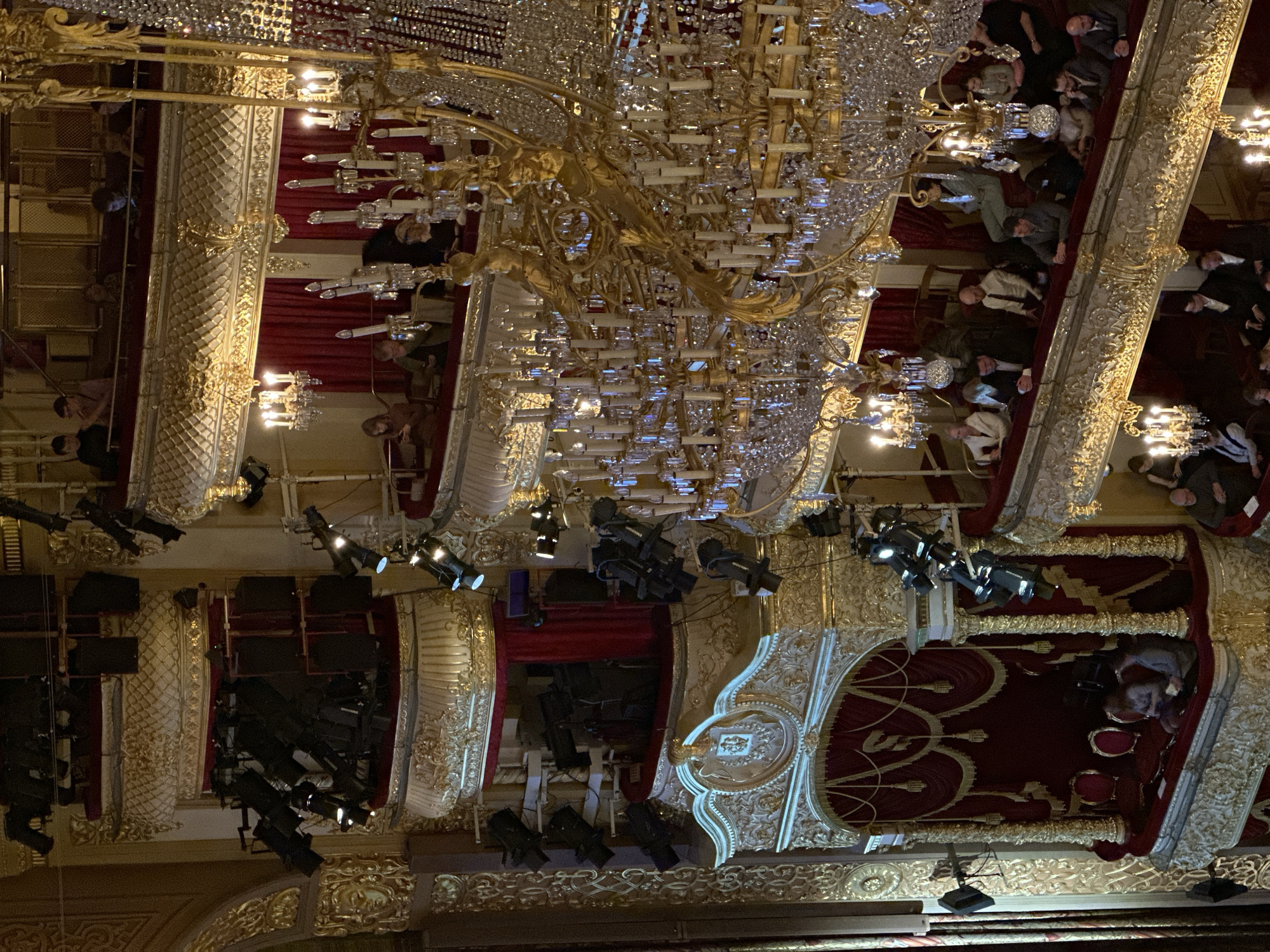 The image captures the opulent interior of a grand theater or opera house, filled with an audience. The dominant feature is an enormous, multi-tiered chandelier adorned with intricate golden scrollwork and countless crystal prisms, hanging from the ceiling.
The venue is richly decorated in a classical style, featuring gilded stucco, ornate carvings, and red velvet drapes and upholstery. Multiple levels of balconies and private boxes line the walls, all elaborately decorated. Many audience members are visible, seated in these boxes and balconies, looking towards an unseen stage. Most individuals are wearing face masks, indicating the time period of the photograph.
Numerous modern stage lights and spotlights are mounted on scaffolding or rigging, positioned throughout the upper areas of the theater, suggesting a live performance is either in progress or about to begin. These lights are pointed towards what would be the stage area, illuminating the space with artificial light. The atmosphere appears formal and attentive.
The scene depicts an event taking place inside, likely an evening performance. The architectural grandeur, combined with the presence of an audience and professional lighting equipment, points to a high-profile cultural event. The location context provided is Khimki, Russia. No visible text is discernible in the image.